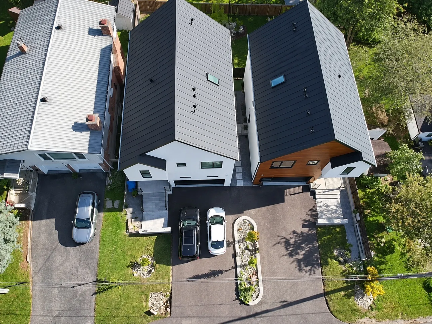 Straight down onto the house and its neighbour: two black metal roofs, two driveways, the planted island at the curb