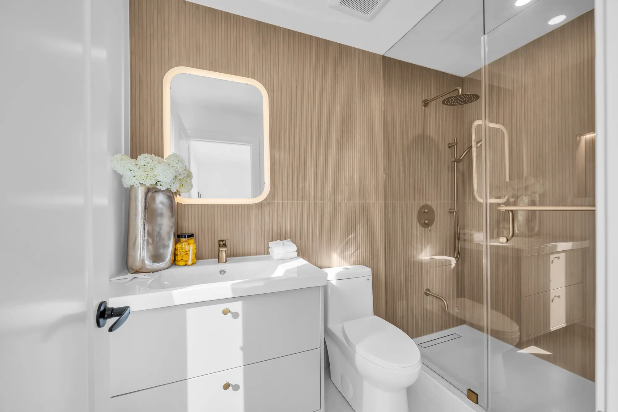 A bathroom with an oak-slat wall: a white vanity with a rounded backlit mirror, a glass shower with a rain head, a toilet