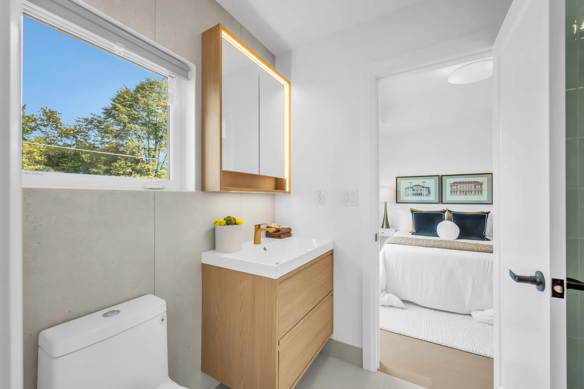 A bathroom with a window: an oak vanity with a white counter, a backlit mirror cabinet, a toilet, the third bedroom through the door