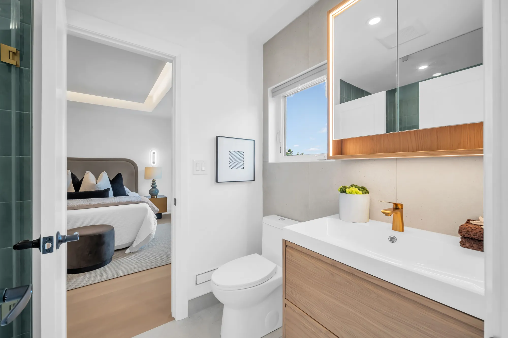 The same bathroom from the other door: the green tile of the shower at the left, the oak vanity, the toilet, a bedroom beyond