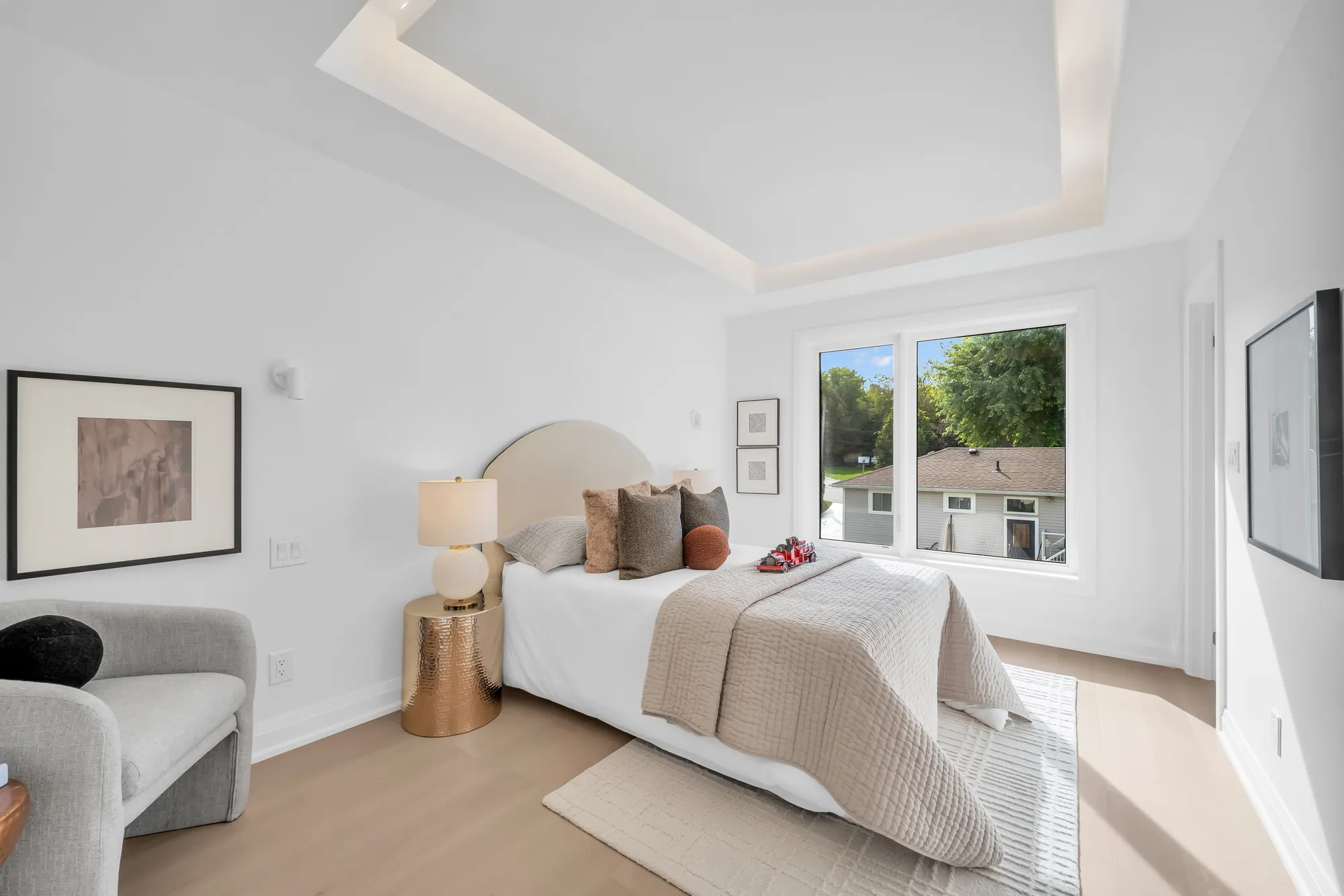 A second bedroom: a bed with an arched cream headboard, a window to the trees, a grey armchair, a brass side table, a tray ceiling
