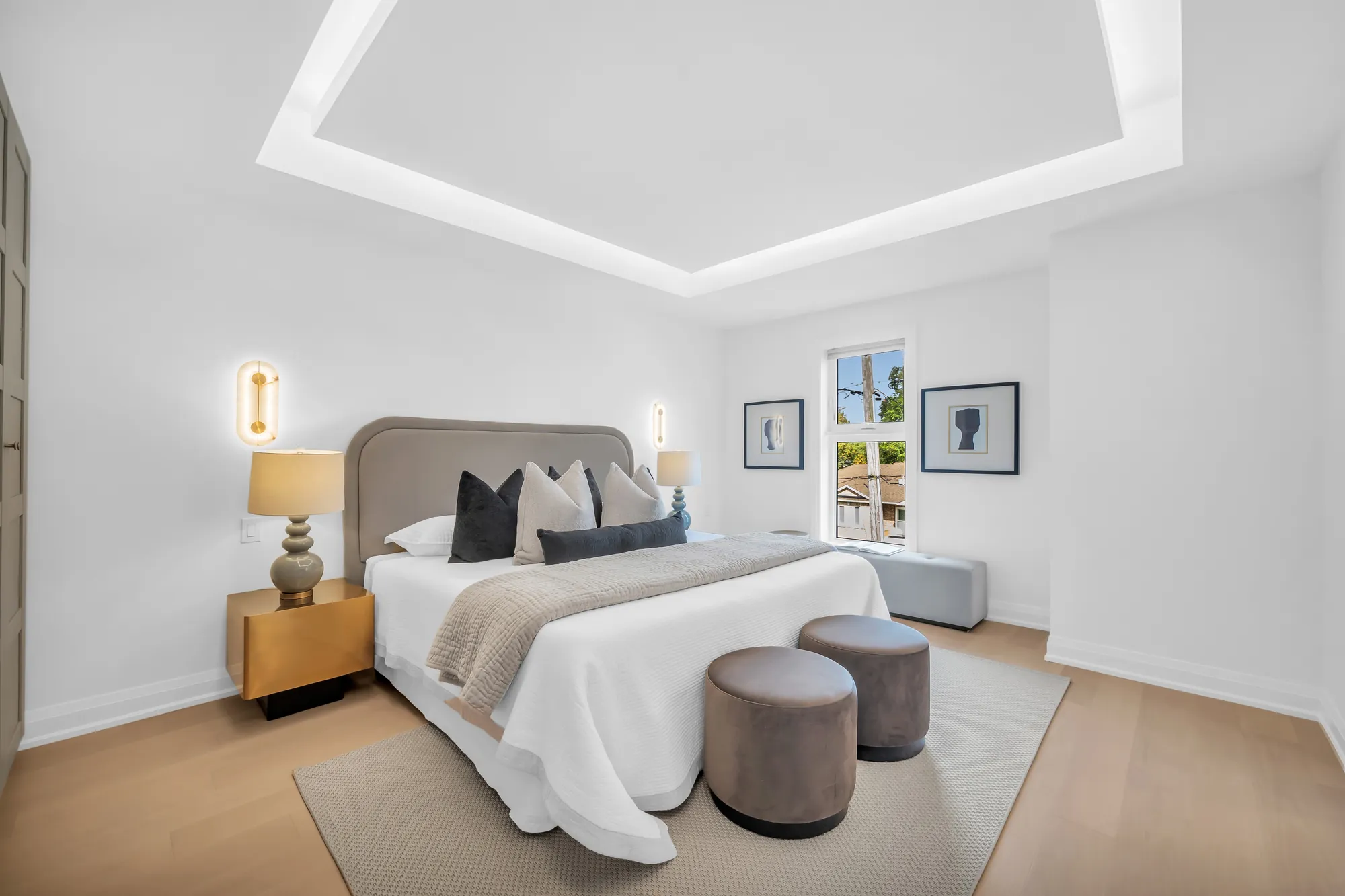 A fourth bedroom: a bed with a grey headboard and black cushions, two brass sconces, framed prints, a window, a tray ceiling