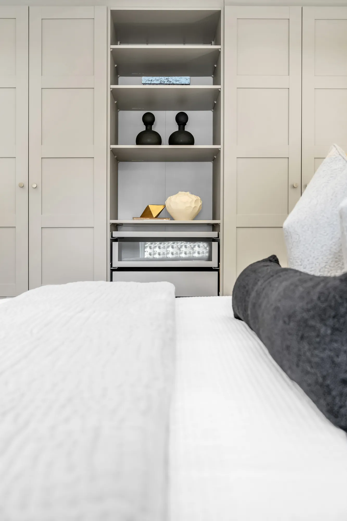 A detail of a bedroom's built-in closet: white shelves with black objects, the bed's dark cushion below