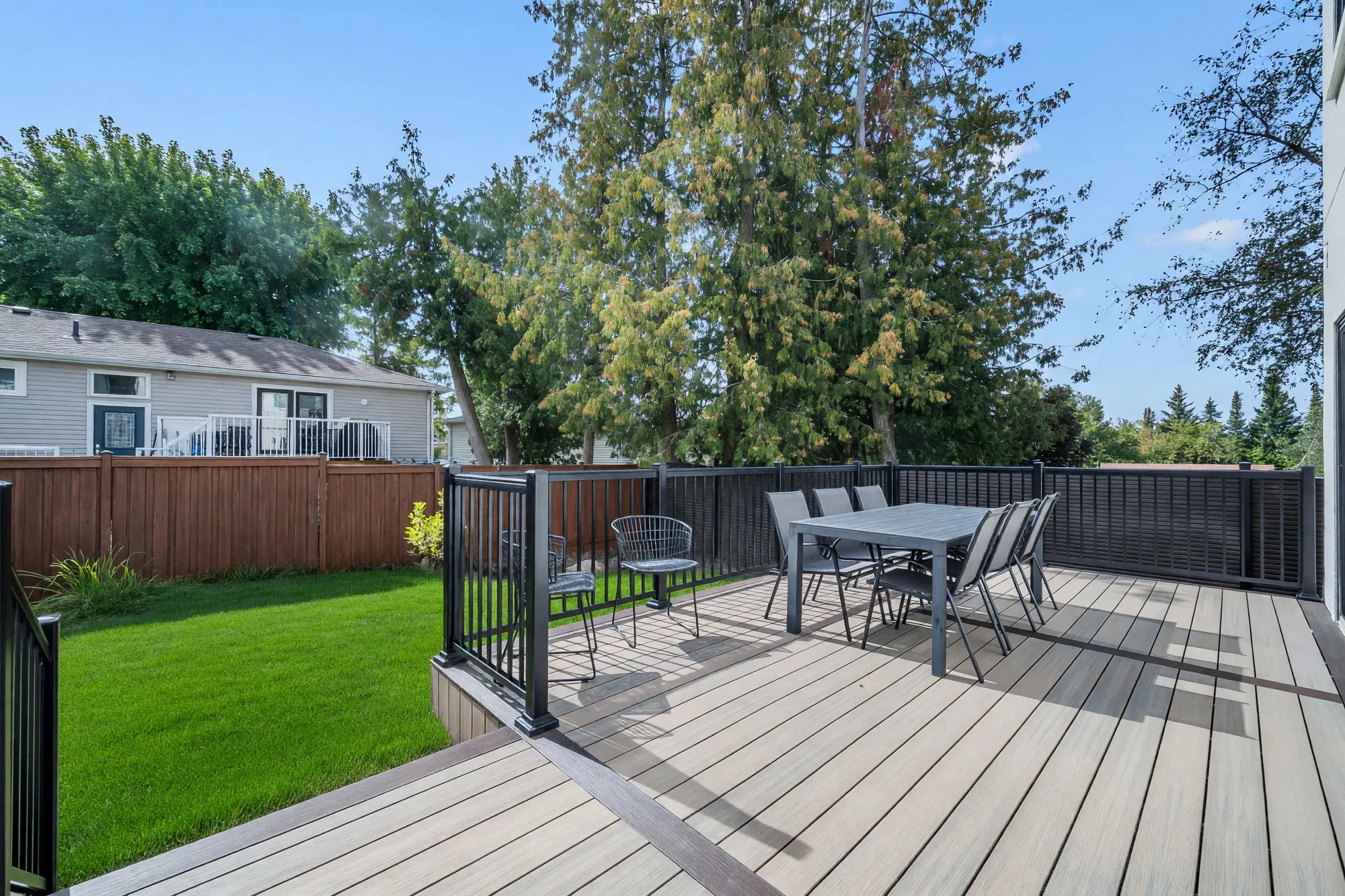 The composite deck: a black railing, a dining table and chairs, the lawn shown green below, a wood fence and the neighbours' trees beyond