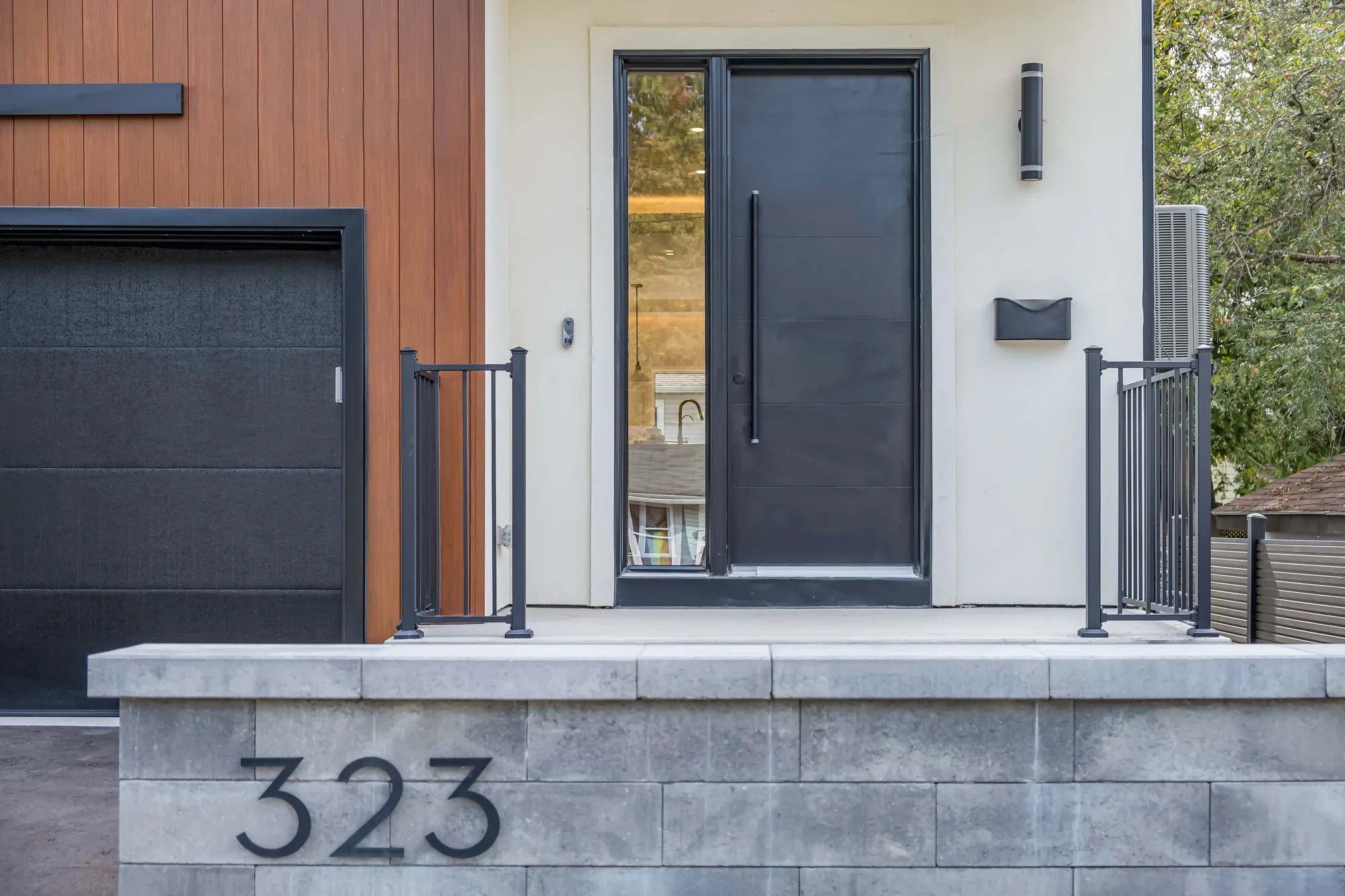 The front door: a black door in a white stucco recess, wood-toned siding beside it, a black railing, the number 323 on the grey block pier below