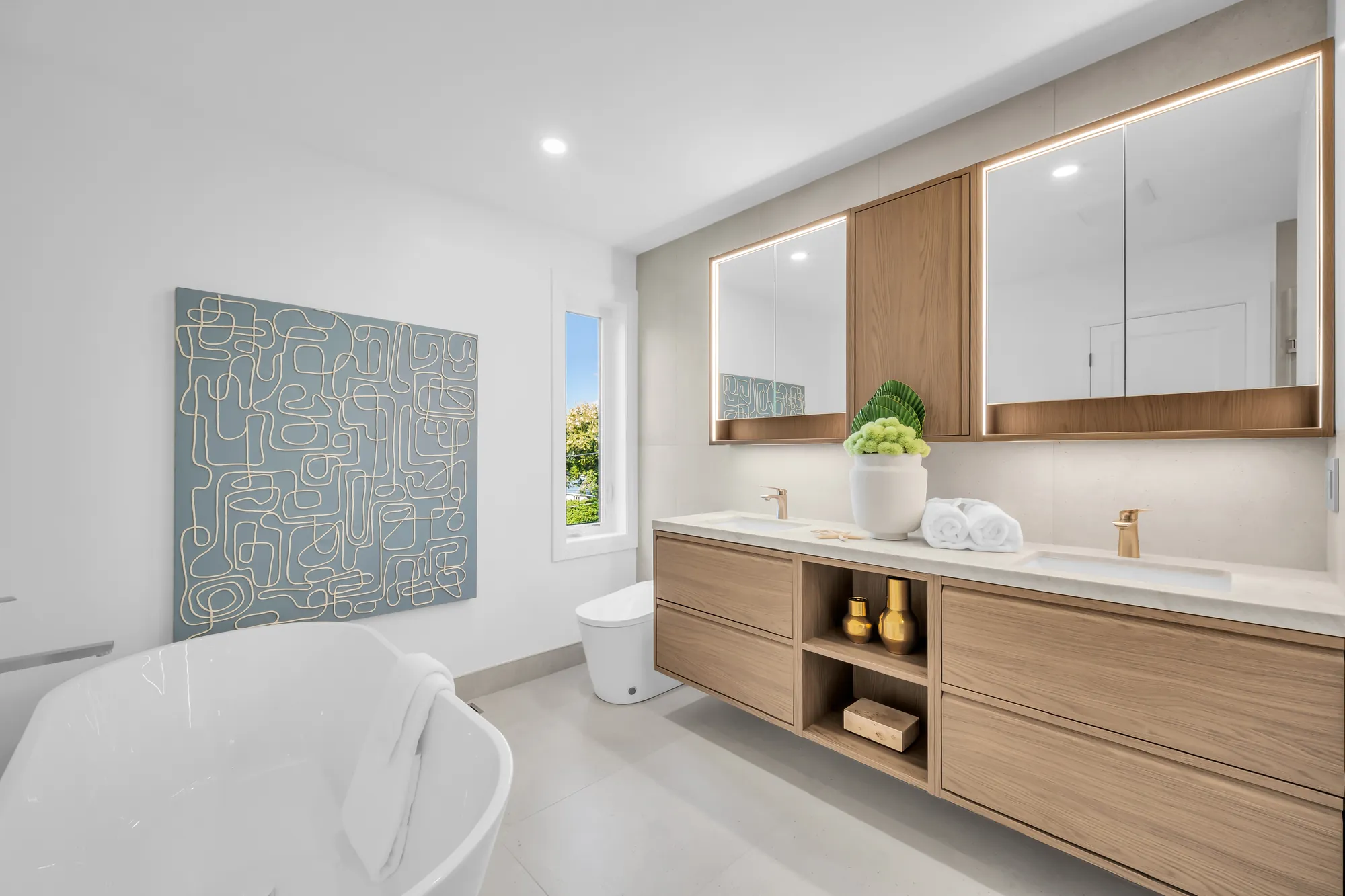 The primary ensuite: a freestanding tub, a long oak double vanity with a white counter and a mirror cabinet, a blue abstract painting, pale tile