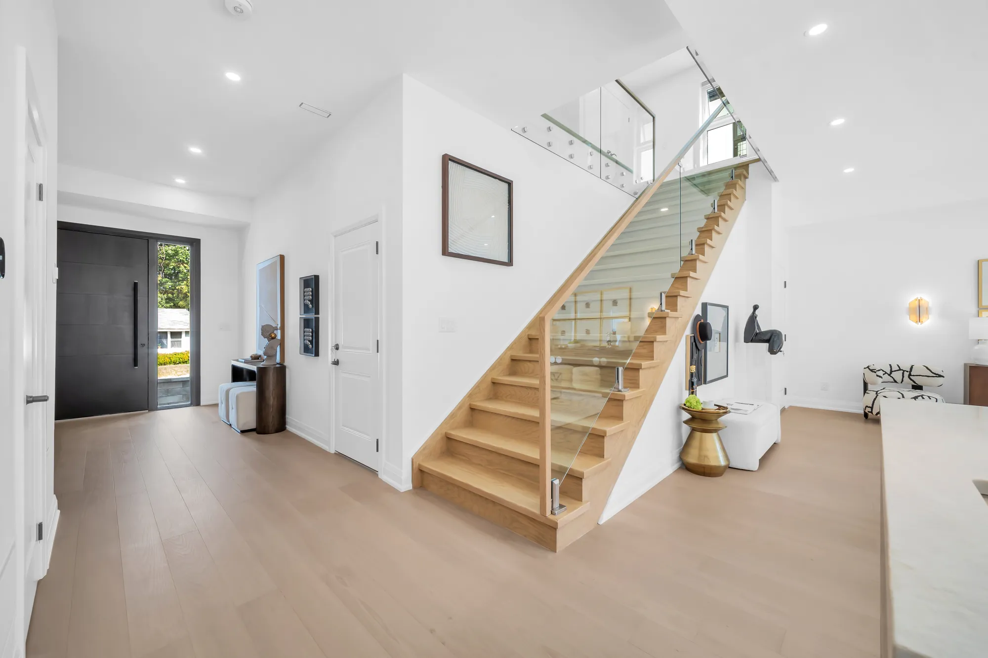 The foyer: the black front door with its sidelight, the oak stair with a glass railing rising to the right, a console and a sculpture under it