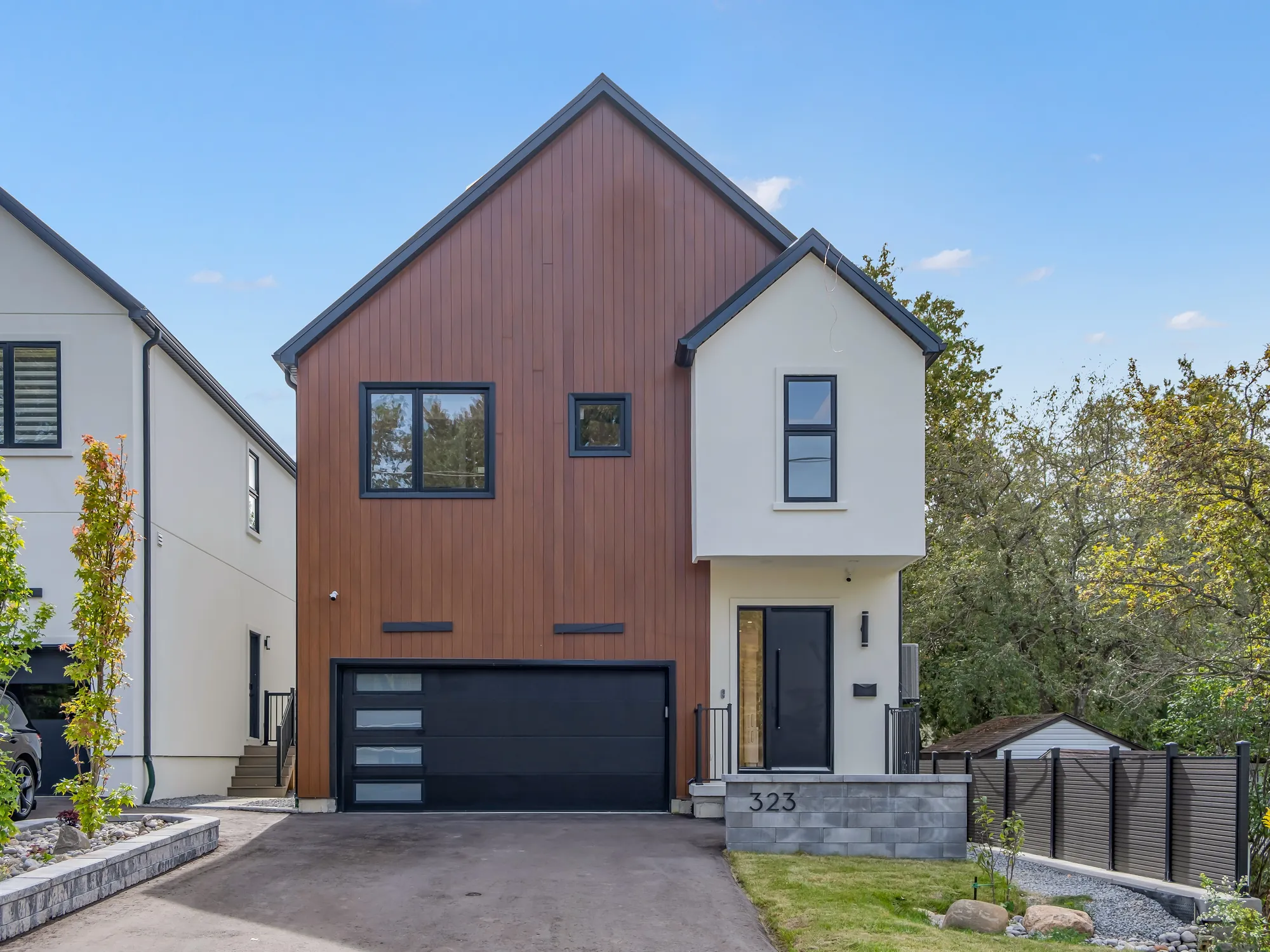 The front of the house by day: a steep gable clad in vertical wood-toned siding over a black garage door with three slit windows, a white stucco wing with a black front door beside it, the number 323 on a stone pier, a blue sky