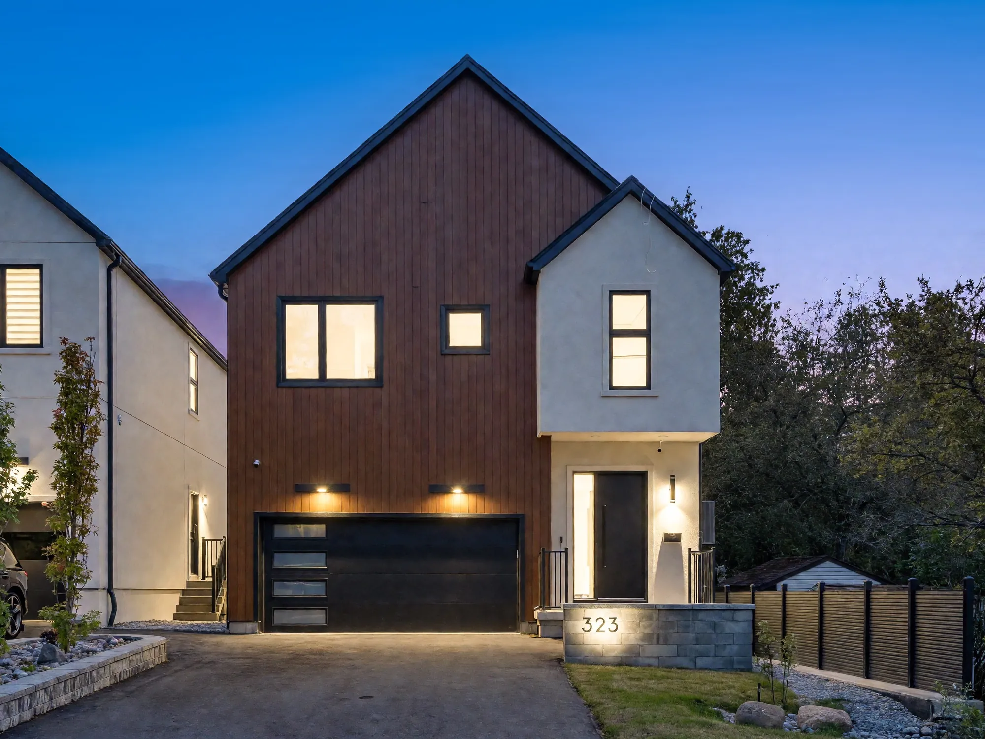 The front of 323 Winnifred Drive at dusk: a steep gable in vertical wood-toned siding over a black garage door, a white stucco wing with the front door beside it, the windows lit against a deep blue sky