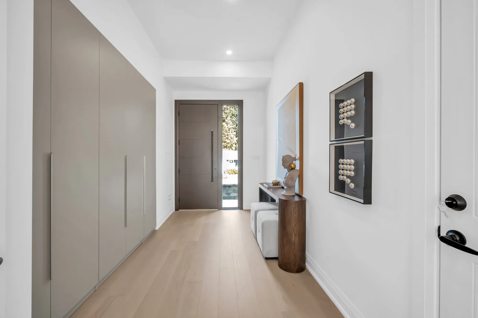 The entry hall: a wall of taupe floor-to-ceiling cabinets, the front door with its sidelight at the end, a round walnut console with a bust under a mirror, two framed pieces on the wall