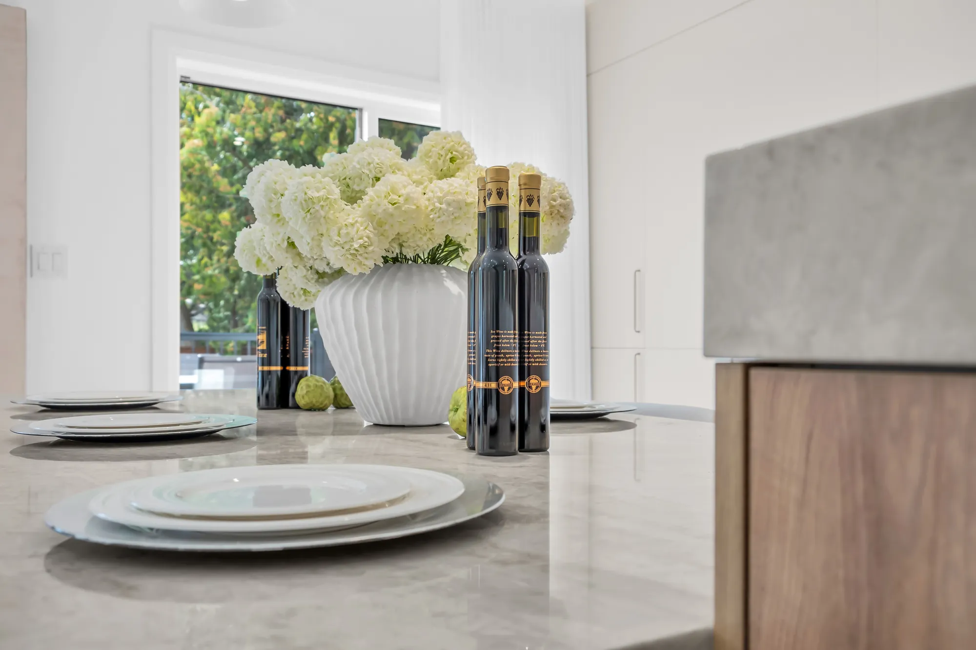 The island's stone counter set with plates, white hydrangeas in a vase, bottles, the window and the trees beyond