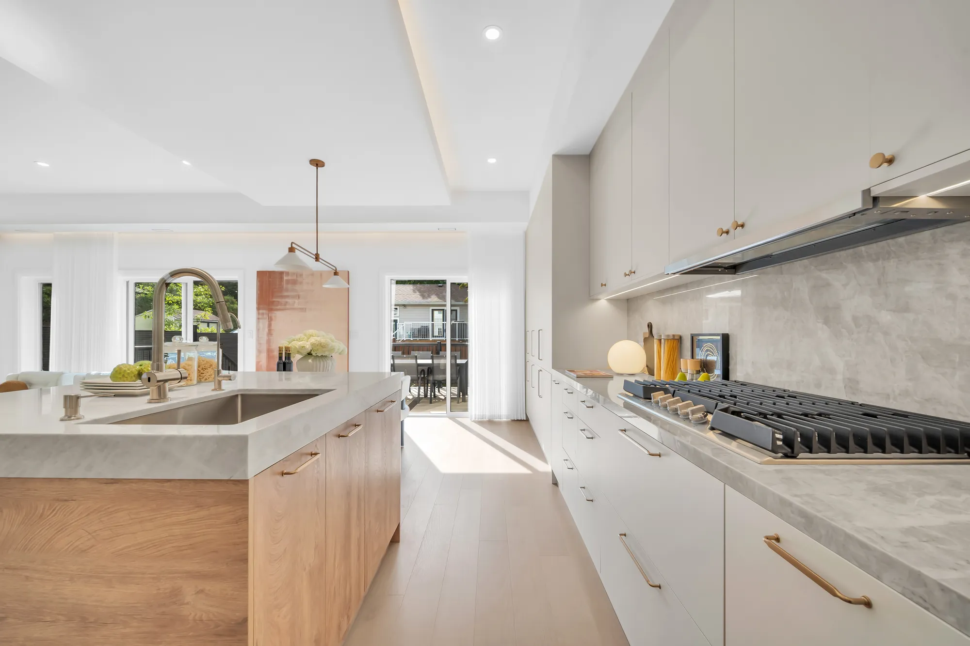 The kitchen: an oak island with a stone waterfall counter and an undermount sink, a run of white cabinets with a stone backsplash, the gas cooktop and the hood, the deck door beyond