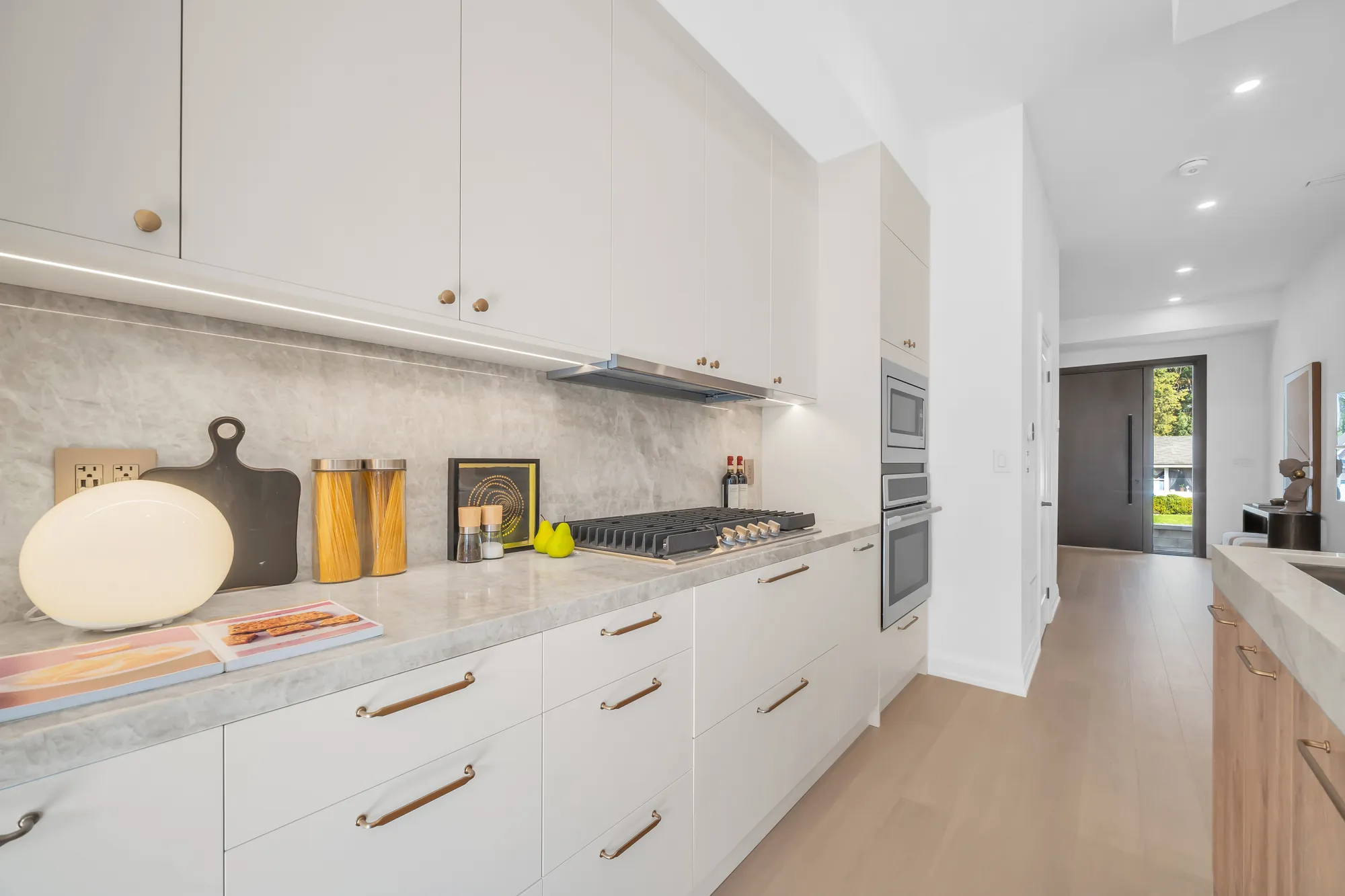 The cooktop wall: white drawers under a stone counter, a stone backsplash, a gas cooktop, a wall oven and a microwave in a tall cabinet, the front hall beyond