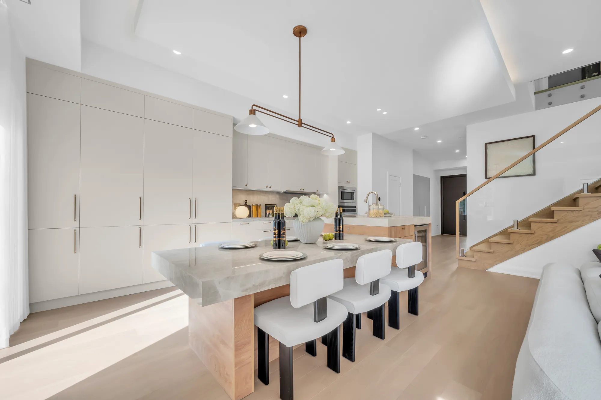 The kitchen from the side: the island with its stools under a pendant, the wall of white cabinets, the stair