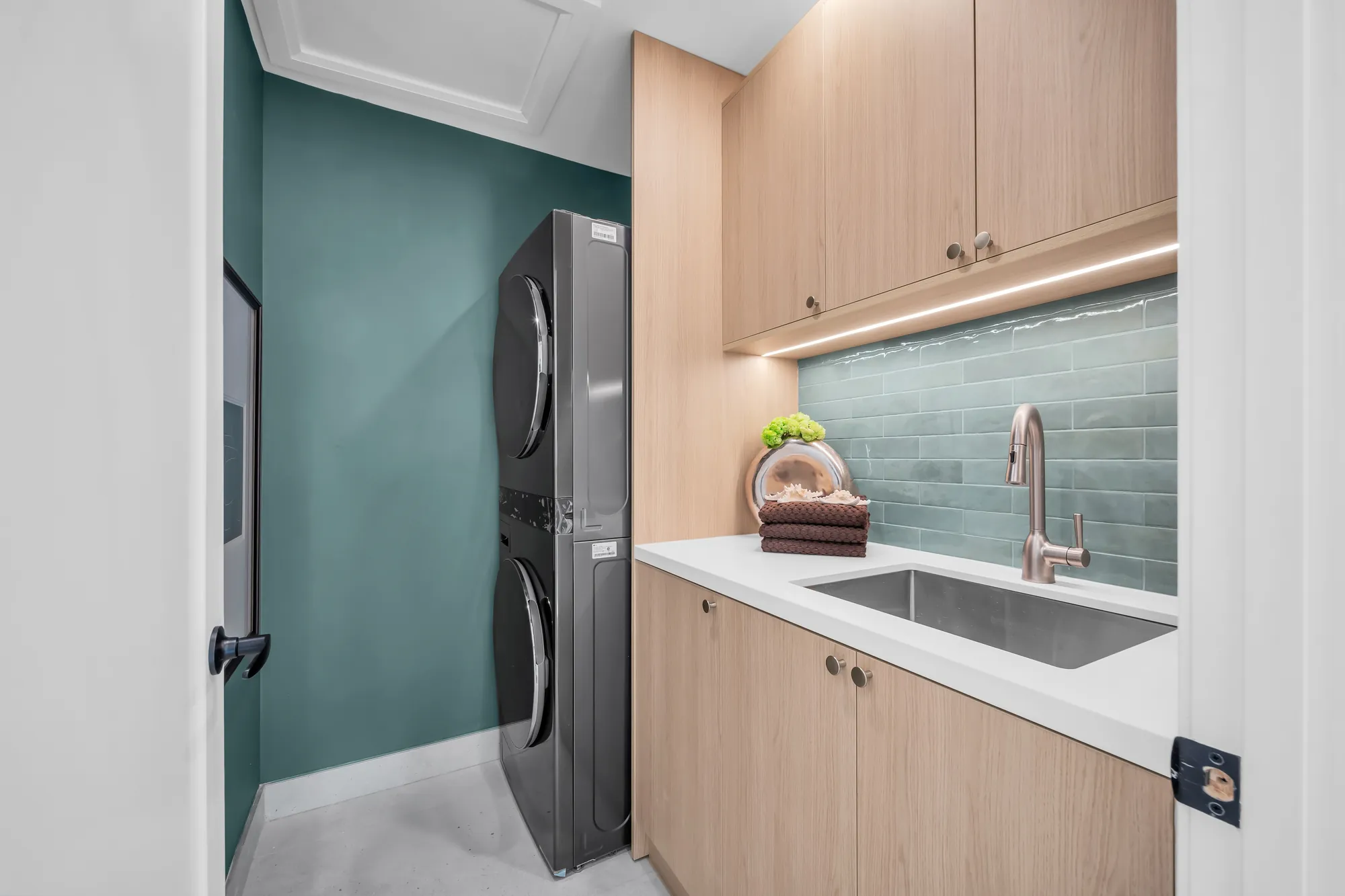 The laundry upstairs: a stacked washer and dryer in a dark green closet, oak cabinets with a white counter and a sink, a green tile backsplash