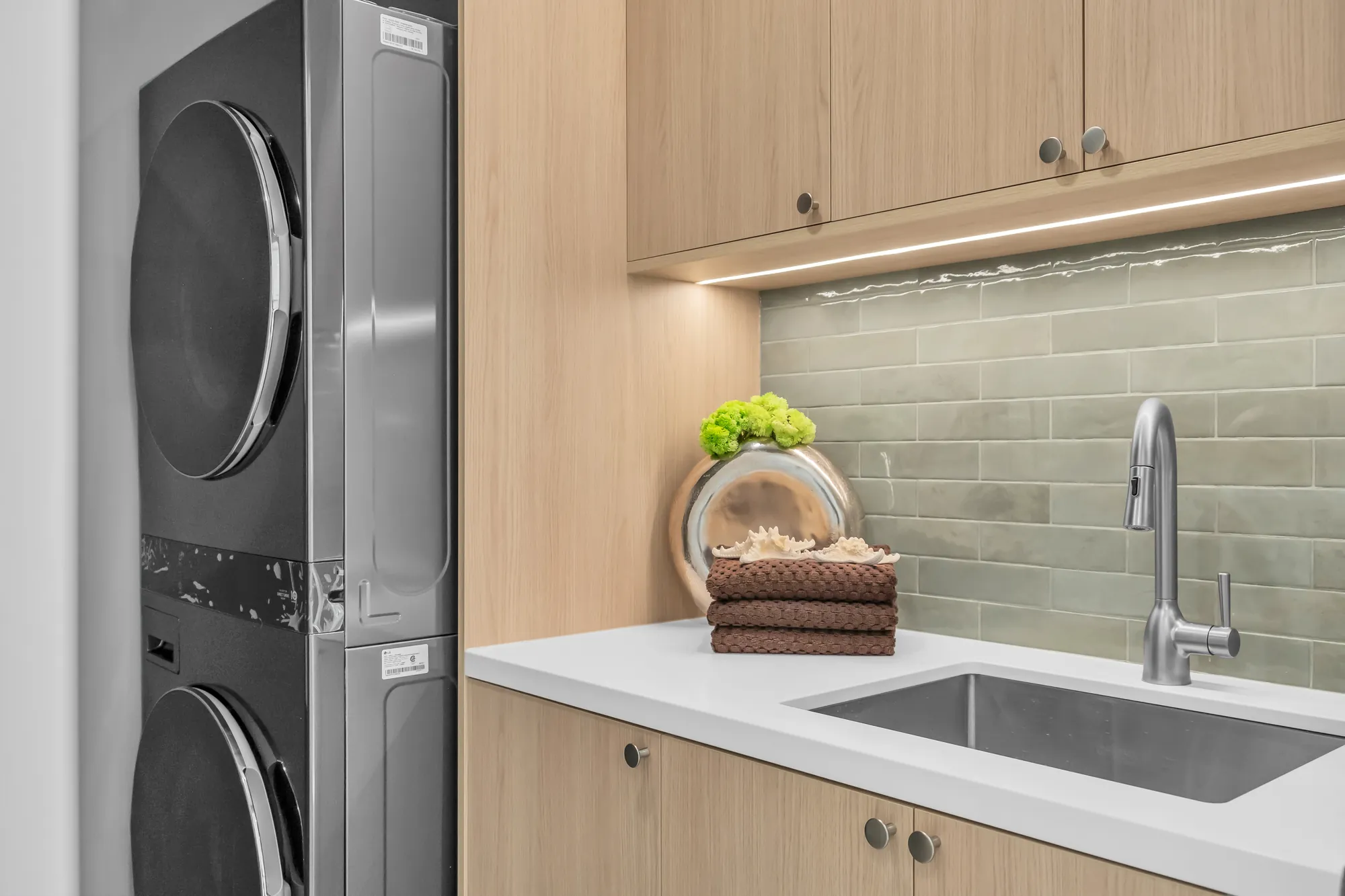 A detail of the laundry: the stacked washer and dryer, the oak cabinet with its lit shelf, the green tile, the sink