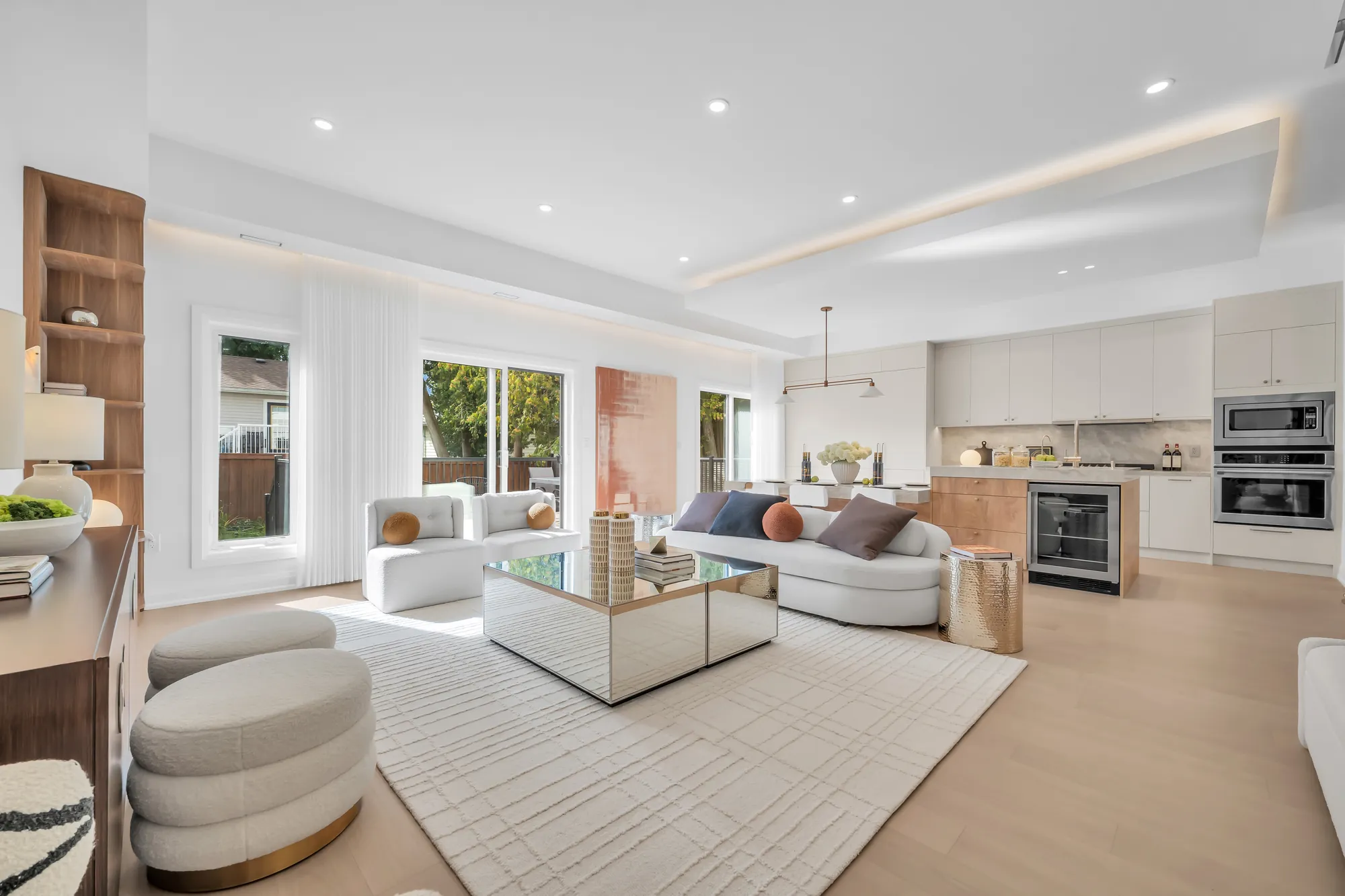 The living room: two white curved sofas around a mirrored coffee table on a pale rug, a walnut console with a grid of framed prints on the wall, sliding doors to the deck at the left, the tray ceiling above