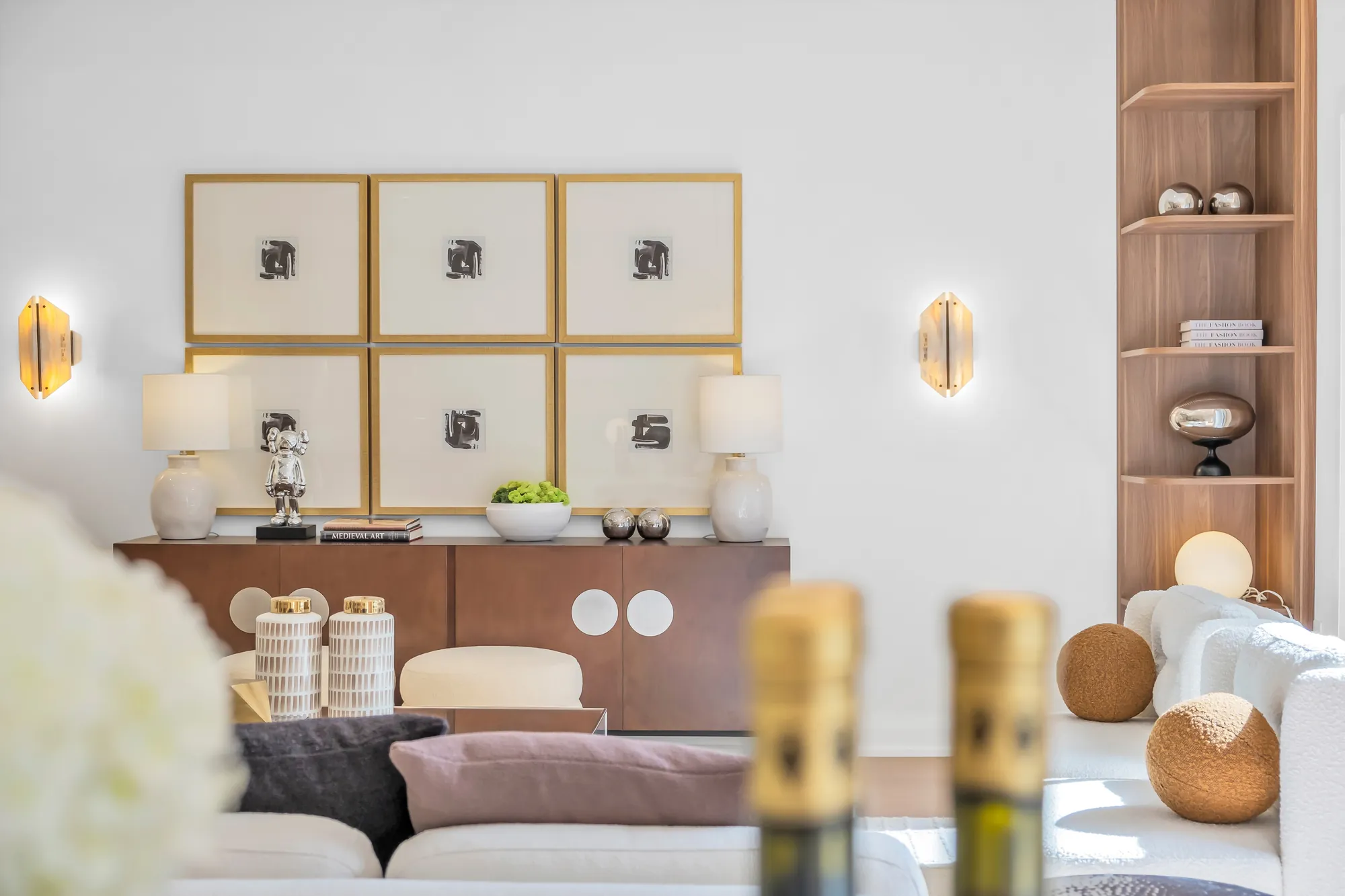 The walnut console: a grid of six framed prints above it, sconces either side, vases and books on top, bottles and hydrangeas in the foreground