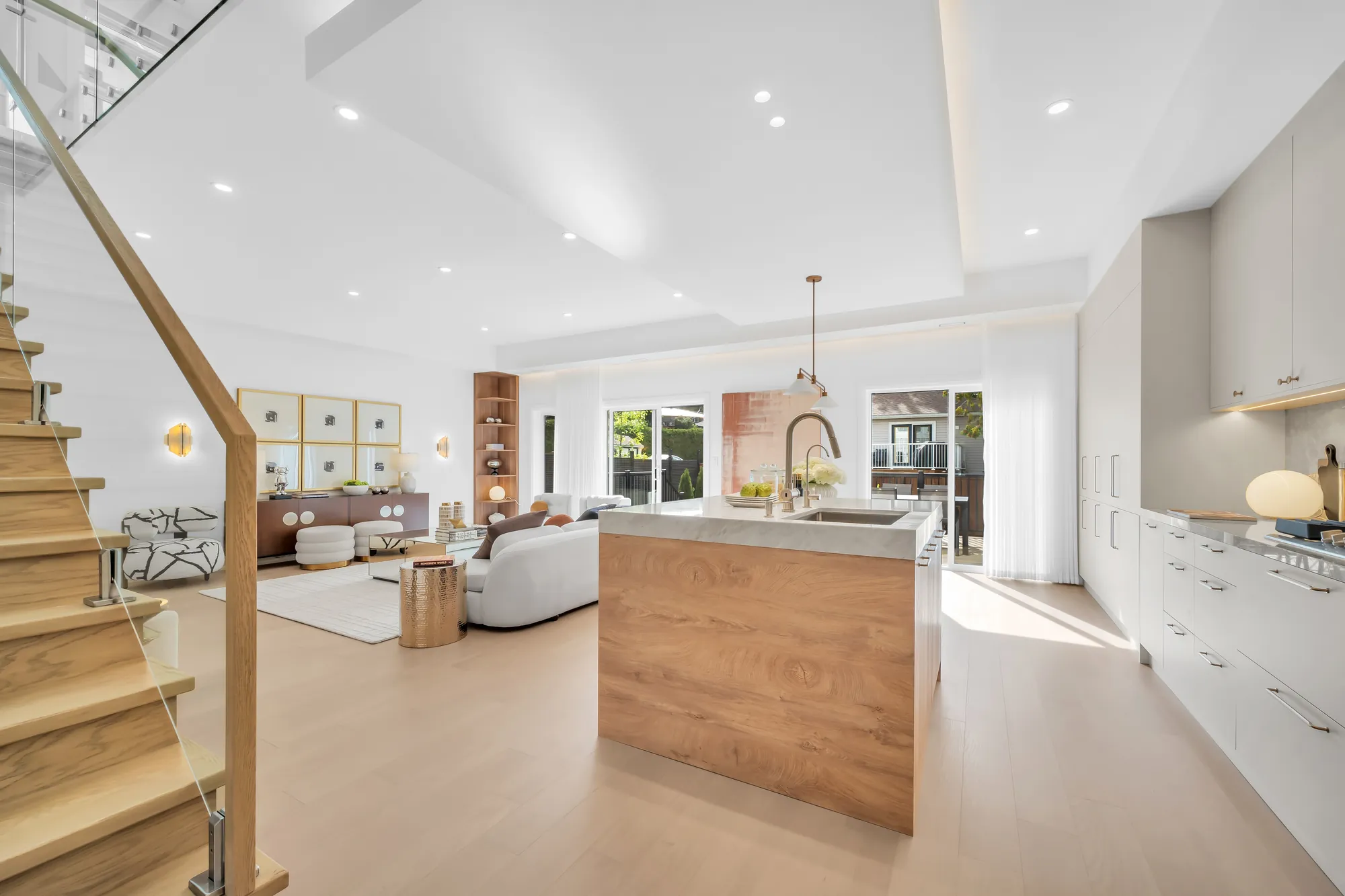 The main floor from the stair: pale oak floors under a recessed tray ceiling, the living room's white sofas at the left, the kitchen island and the white cabinets at the right, the deck doors at the far end