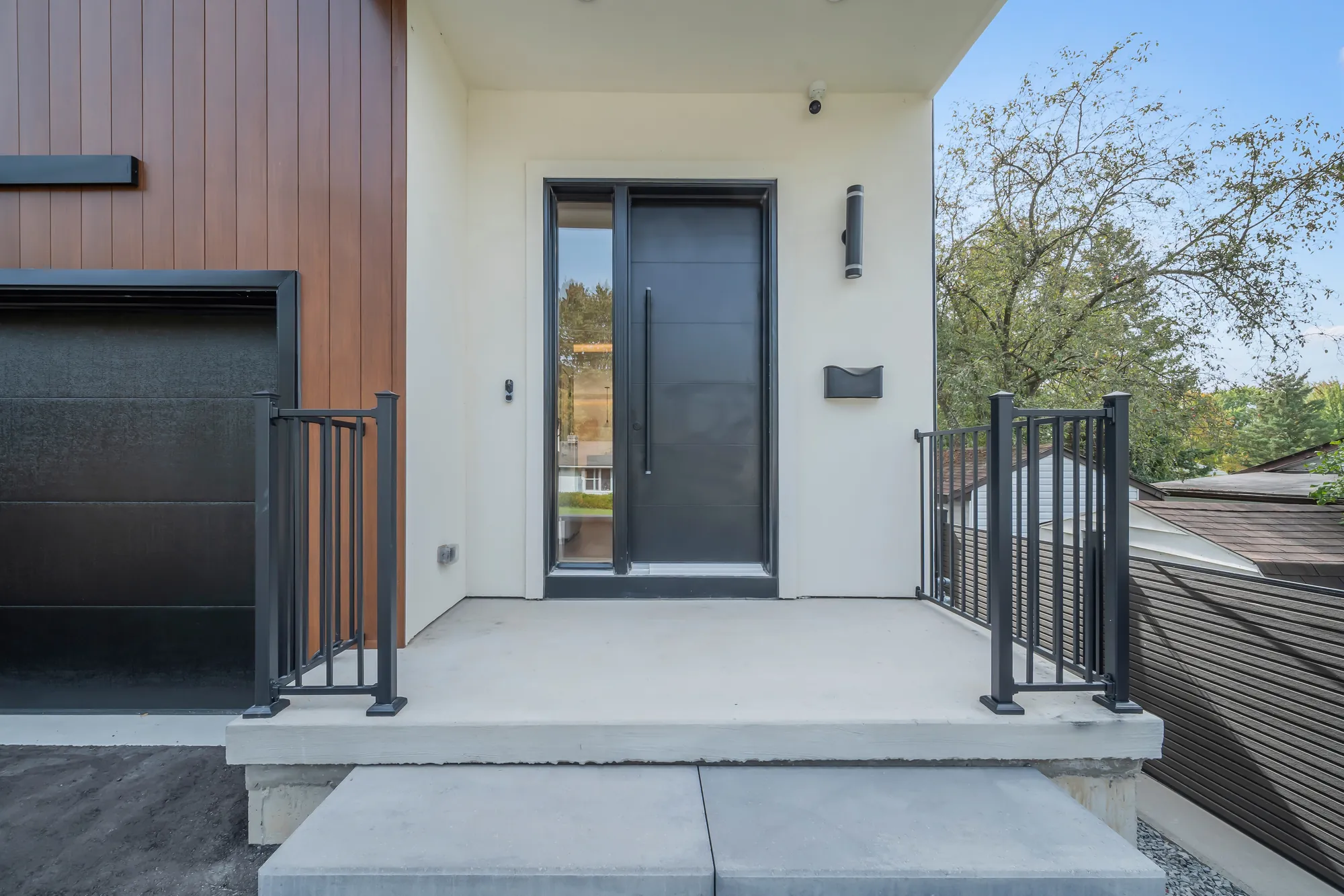 The front step from beside the garage: the black door with its glass sidelight, the concrete landing, black railings, the wood-clad wall