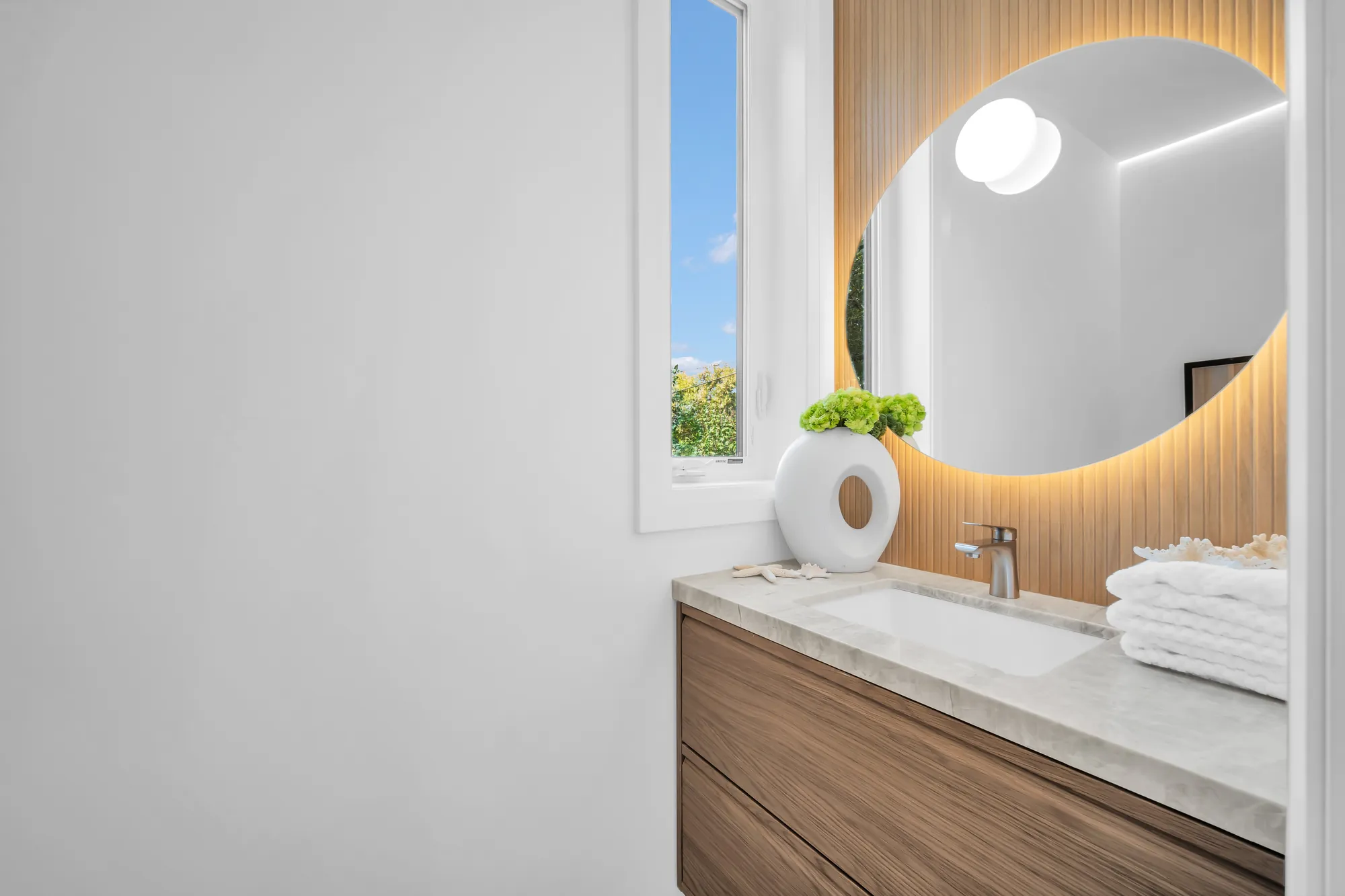 The powder room: an oak vanity with a stone counter, a round backlit mirror, a window, oak slats