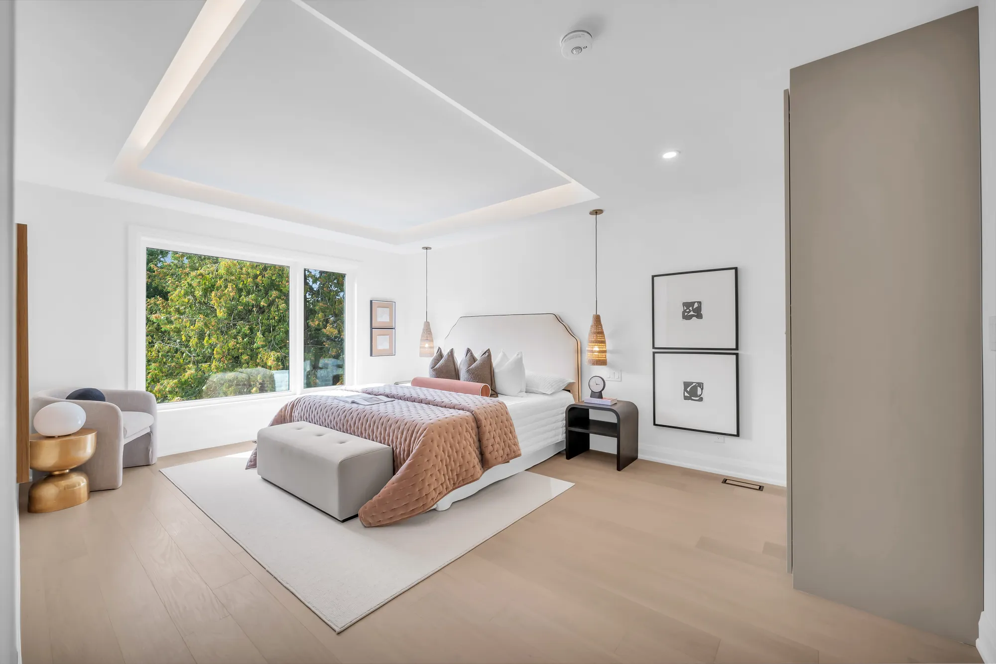 The primary bedroom: a bed with a curved white headboard and a pink throw under two rattan pendants, a recessed tray ceiling, a window to the trees, a grey armchair, a wall of built-in wardrobes