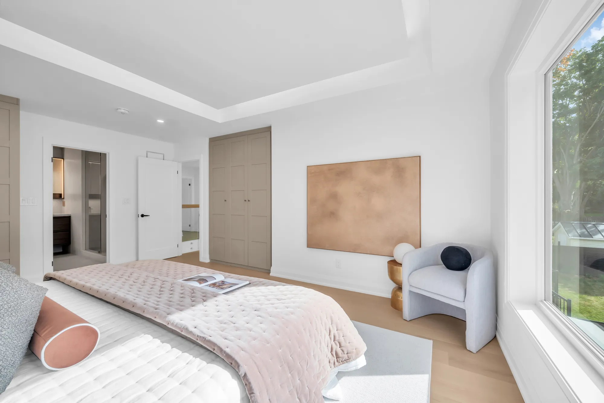 The primary bedroom toward its door: the taupe wardrobe doors, a copper-toned art panel, the grey armchair by the window, the ensuite through the door at the left