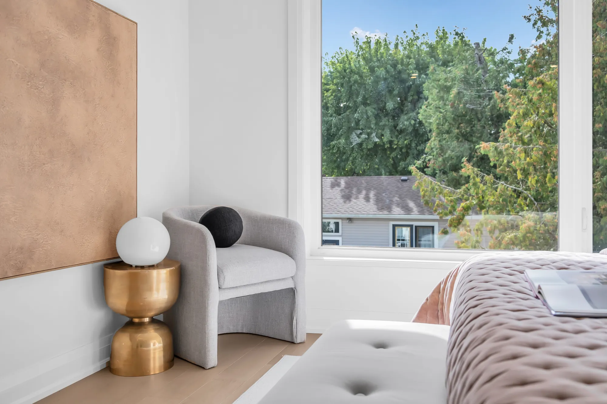 A detail of the primary bedroom: the grey armchair by the window with a brass side table, the pink throw on the bed, the trees outside