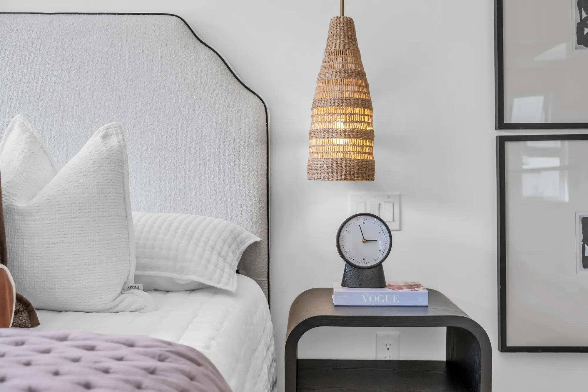 A detail of the primary bedroom: the rattan pendant over a black night table with a clock, the curved headboard, the pink throw