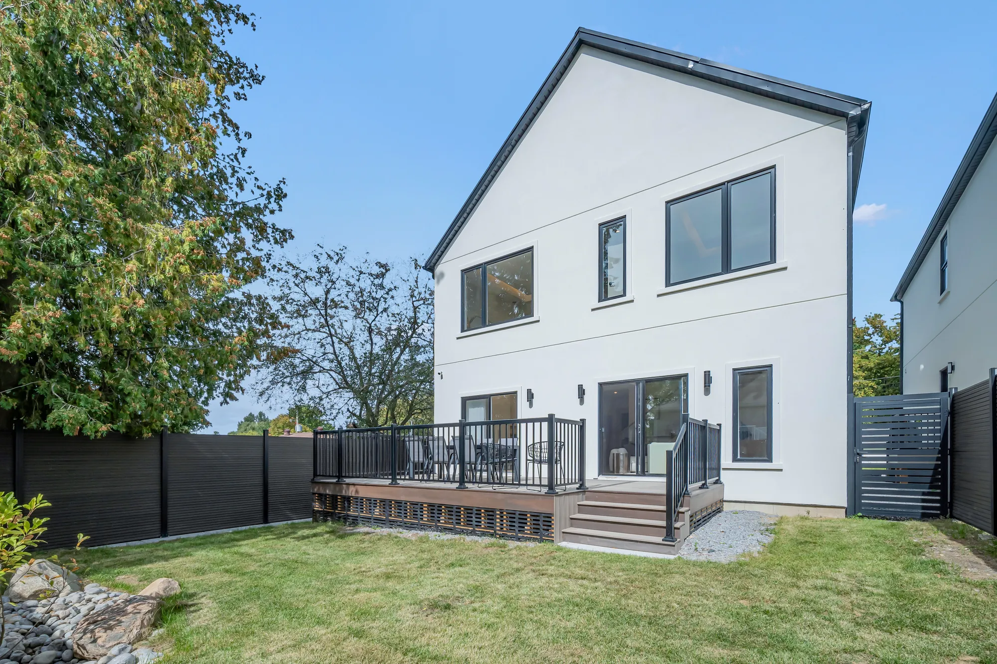 The back of the house from the lawn: a white stucco gable with a black metal roof, sliding doors from the living room onto a composite deck with a black railing, steps down to the lawn, a black fence