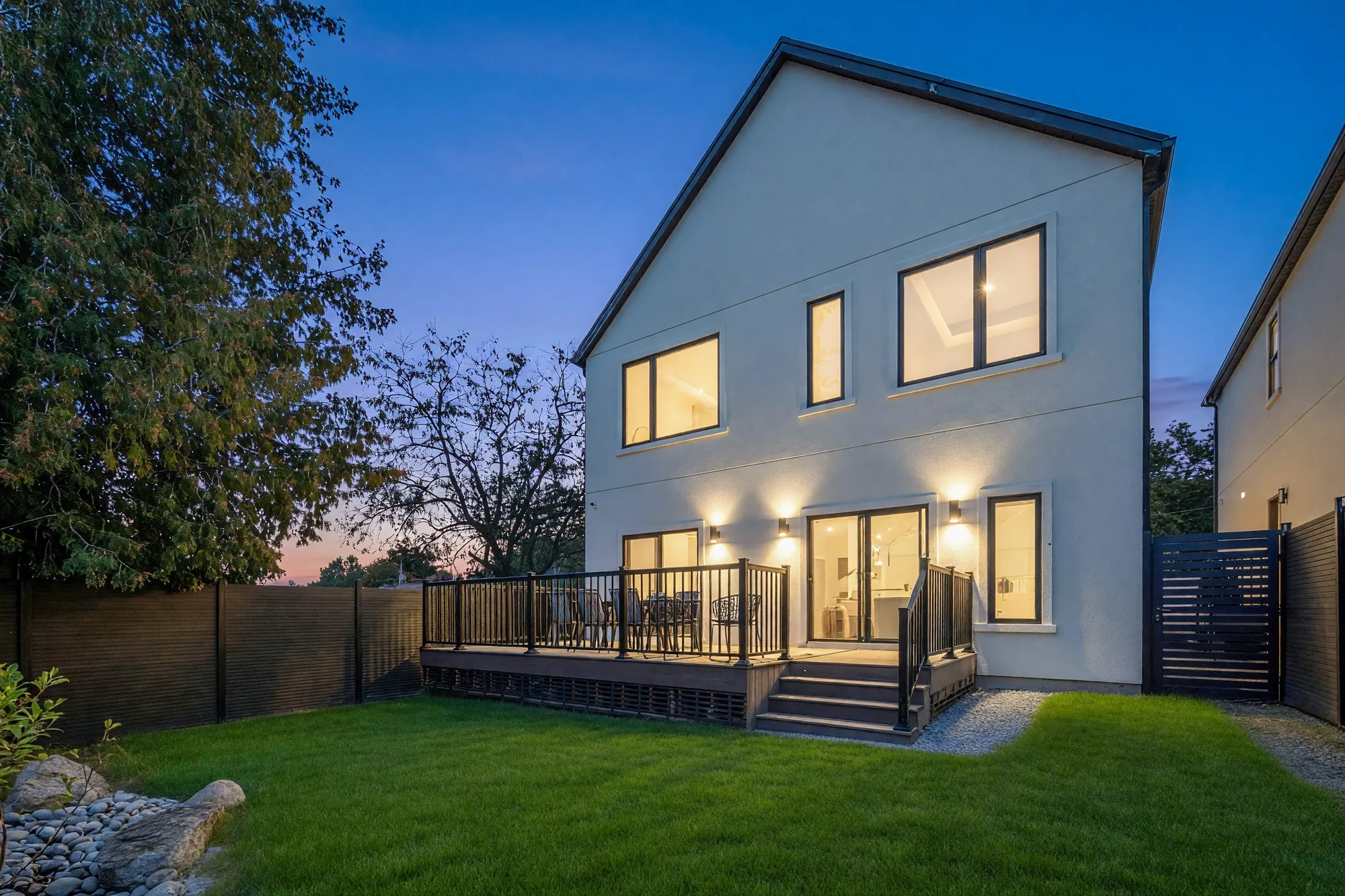 The same view of the back at dusk, the windows and the wall lights lit, the lawn shown green