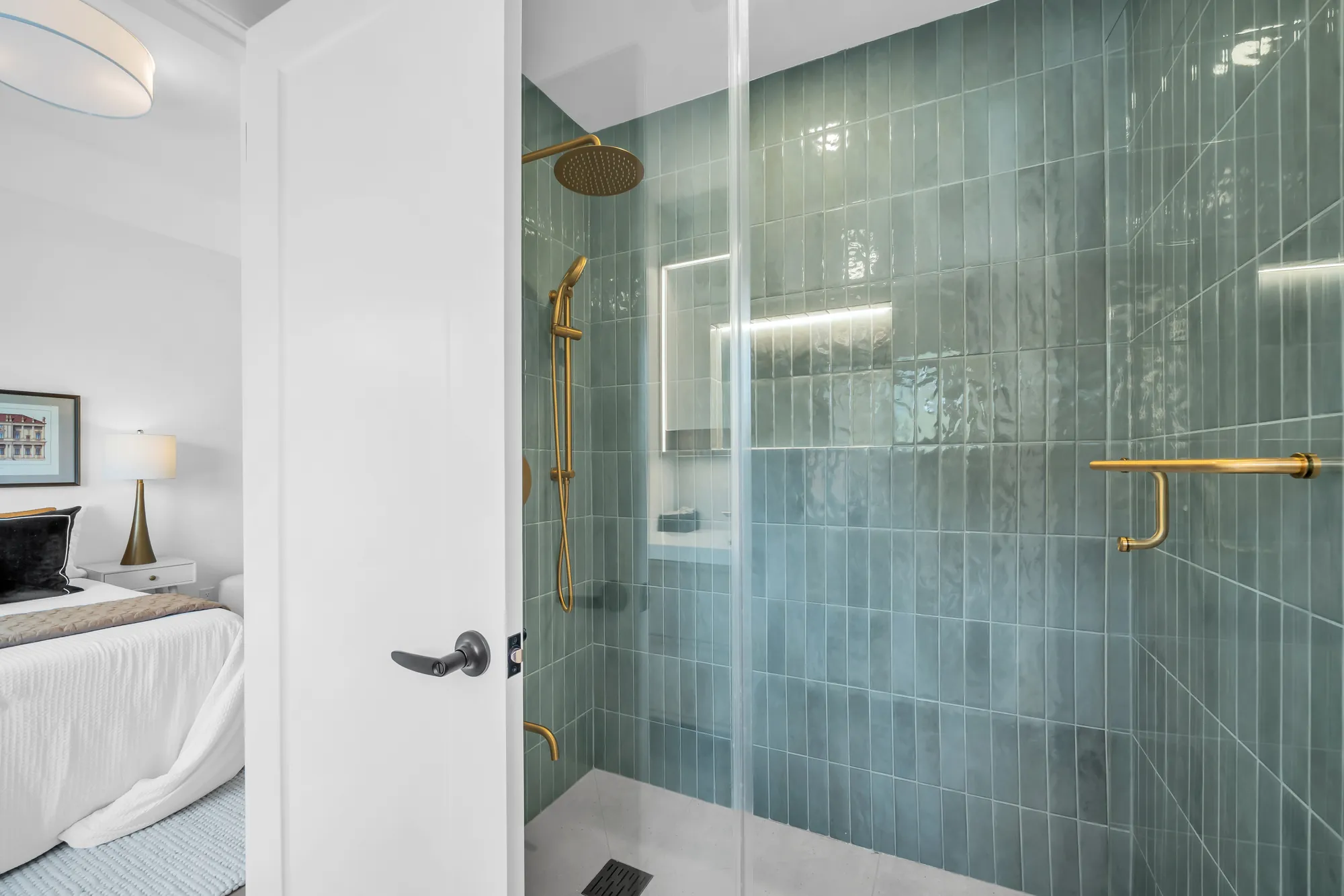 A shower in green tile: a brass rain head and hand shower, a lit niche, a glass door