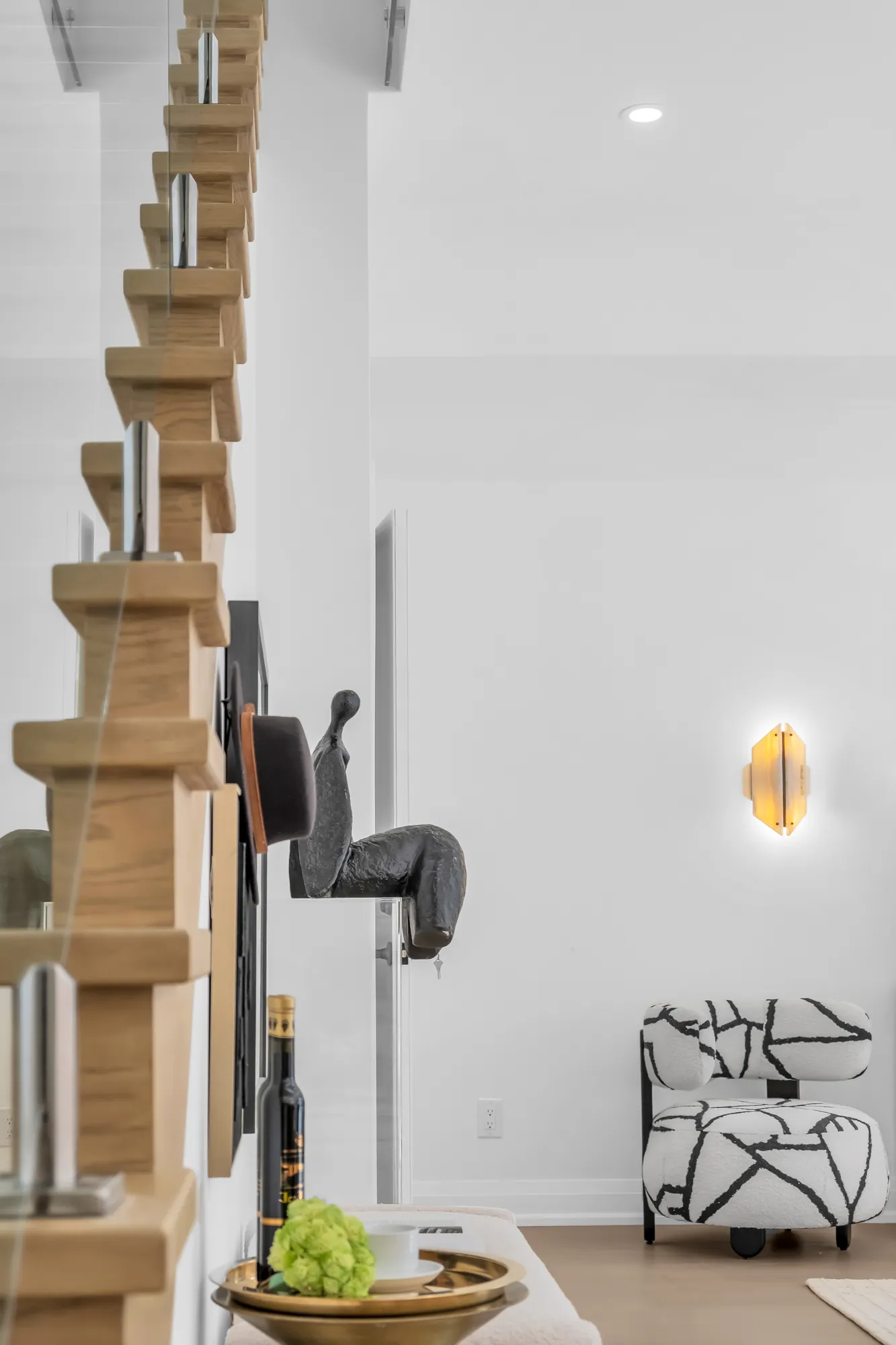 The underside of the stair's oak treads seen through the glass rail from the living room, a black sculpture on the wall, a sconce, a patterned armchair