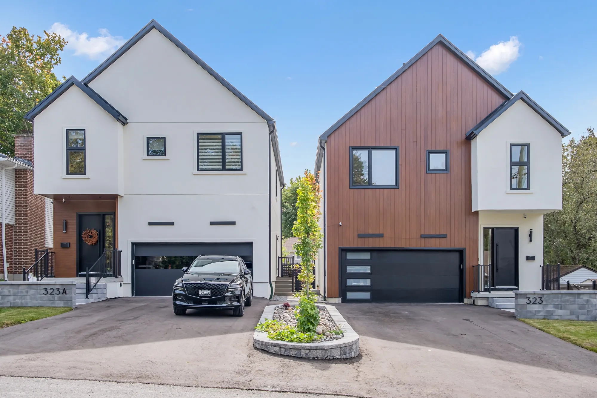 From the street: the house with its wood-clad gable at the right, the house next door at the left, the two driveways between them, a planted island at the curb