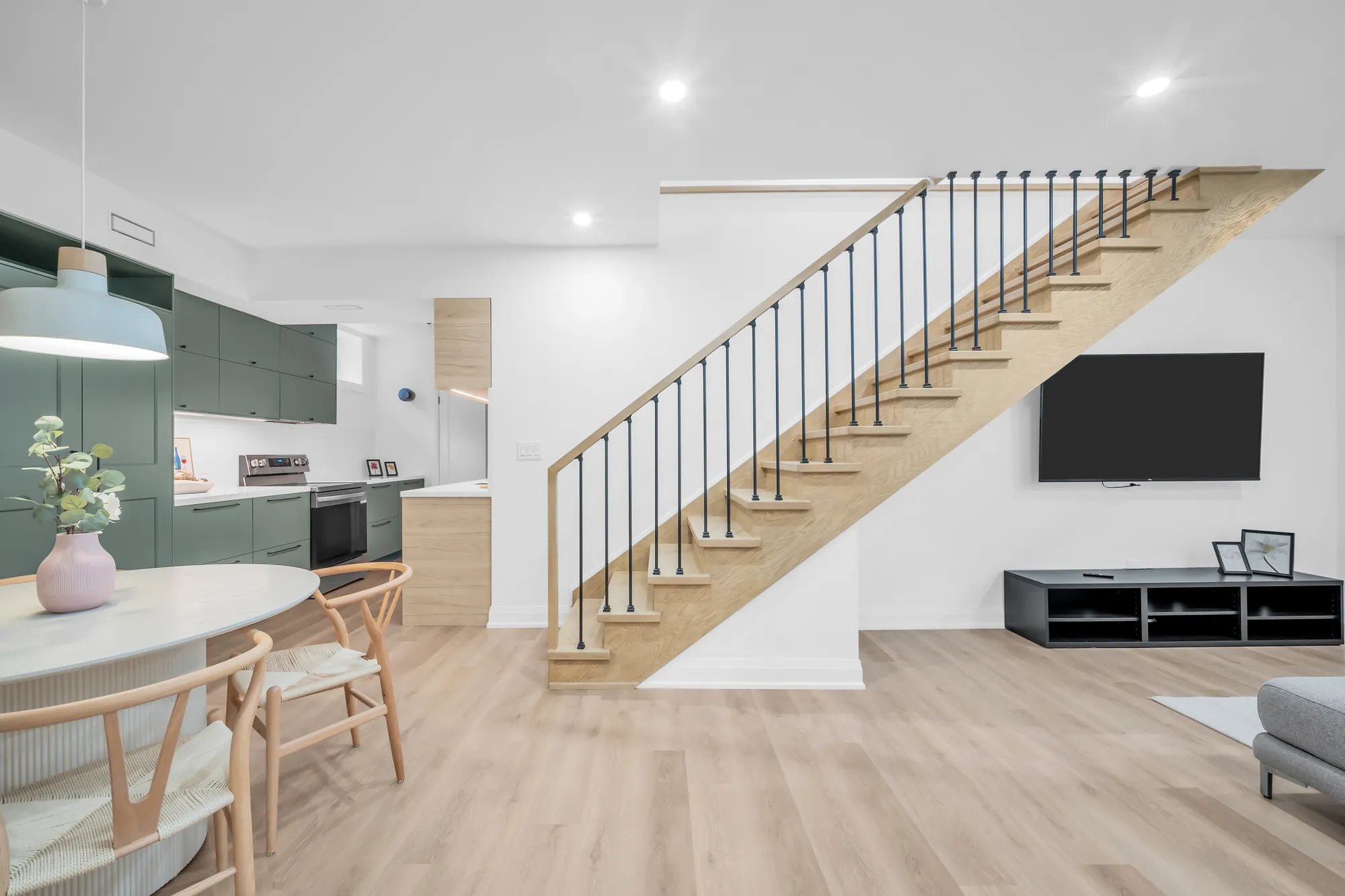 The lower level: a kitchen in sage green cabinets at the left, the stair with black metal spindles, a television on the wall, oak-look floors, a small window