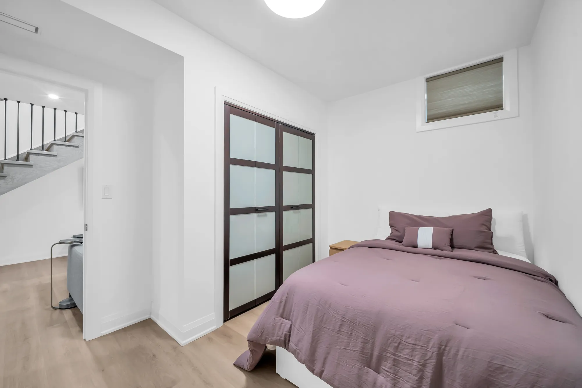A bedroom on the lower level: a bed with a mauve cover, a wood night table, a window at the top of the wall, a sliding-door closet in black frames, the stair through the door