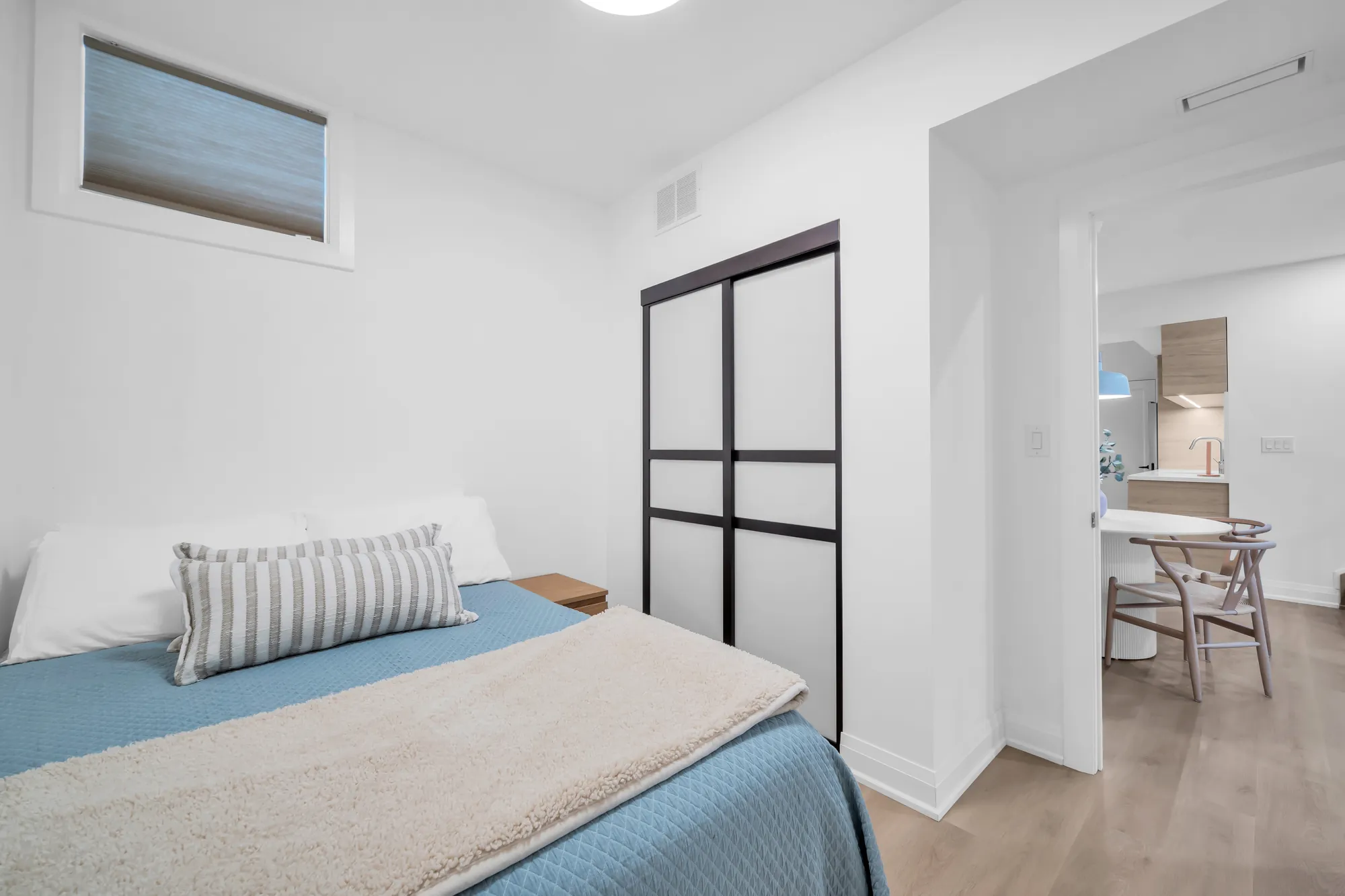 A second bedroom on the lower level: a bed with a blue cover, a sliding-door closet in black frames, a window, the kitchen through the door