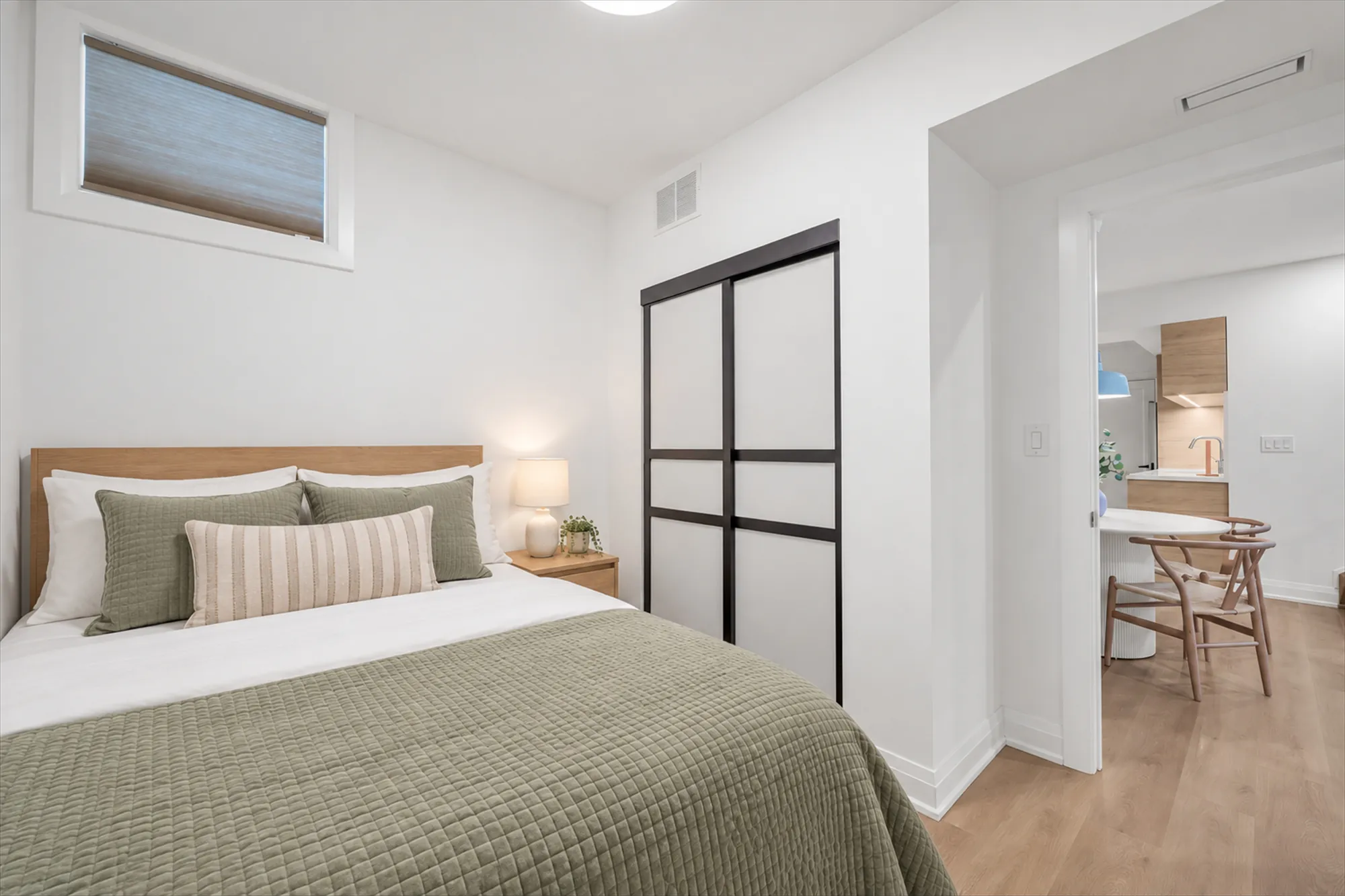 The same second bedroom virtually staged: a bed with green linen and striped cushions, a night table with a lamp, a chair at the table beyond