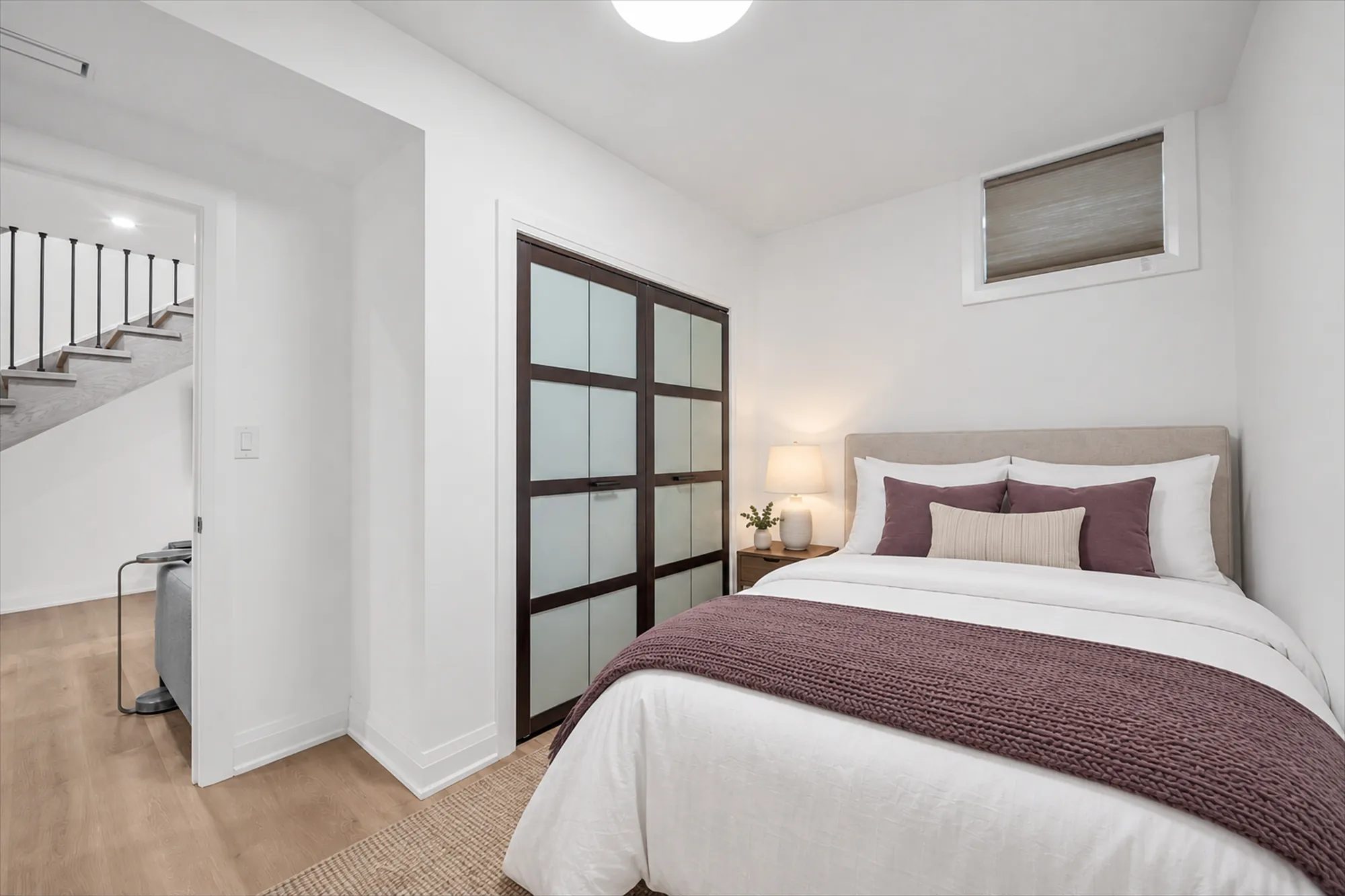 The same bedroom virtually staged: a bed with white linen and a plum throw, a night table with a lamp, a rug and a cushion
