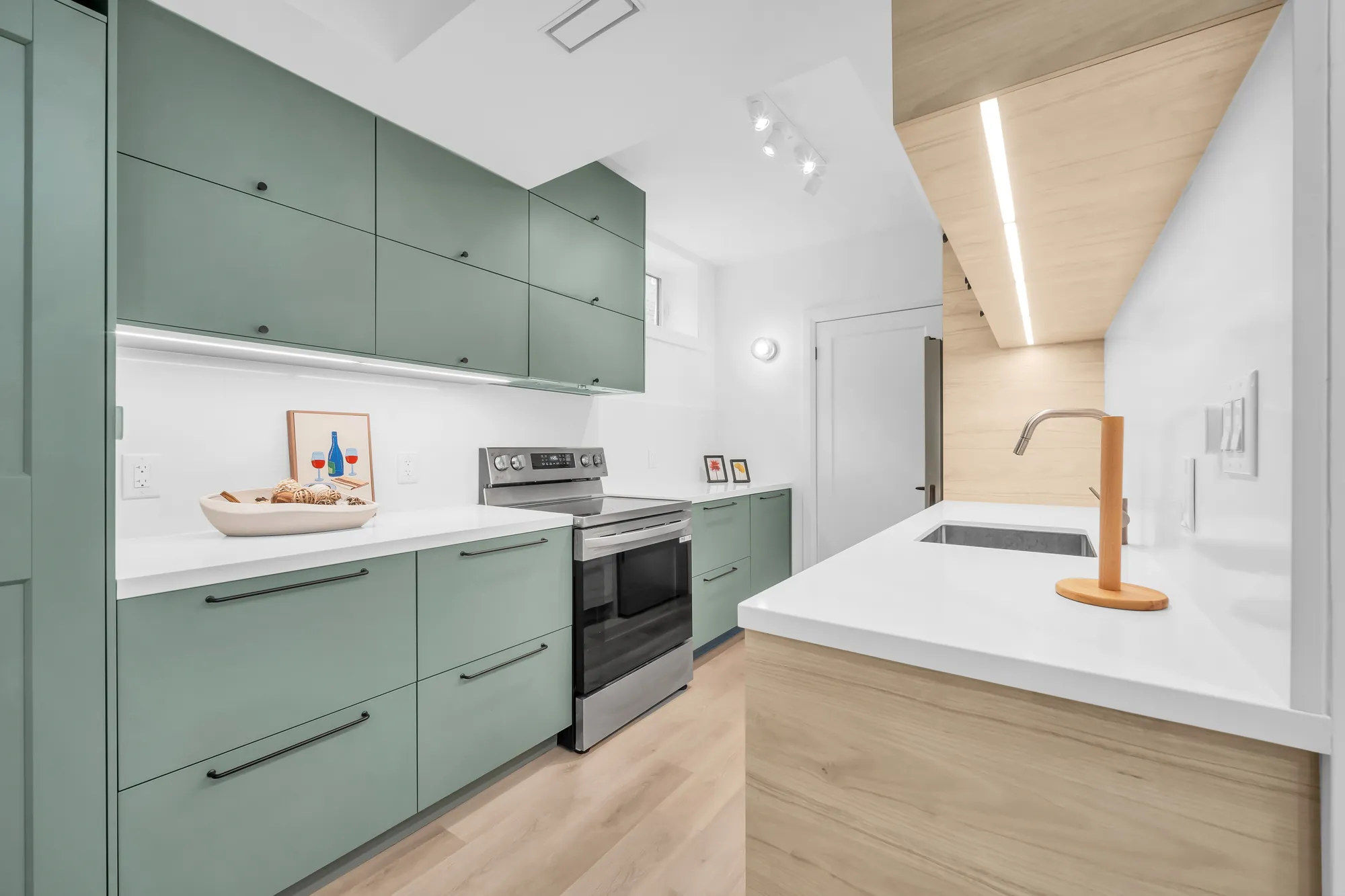 The lower level's kitchen: sage green cabinets, a stainless range, a white counter with an undermount sink, a lit oak upper cabinet