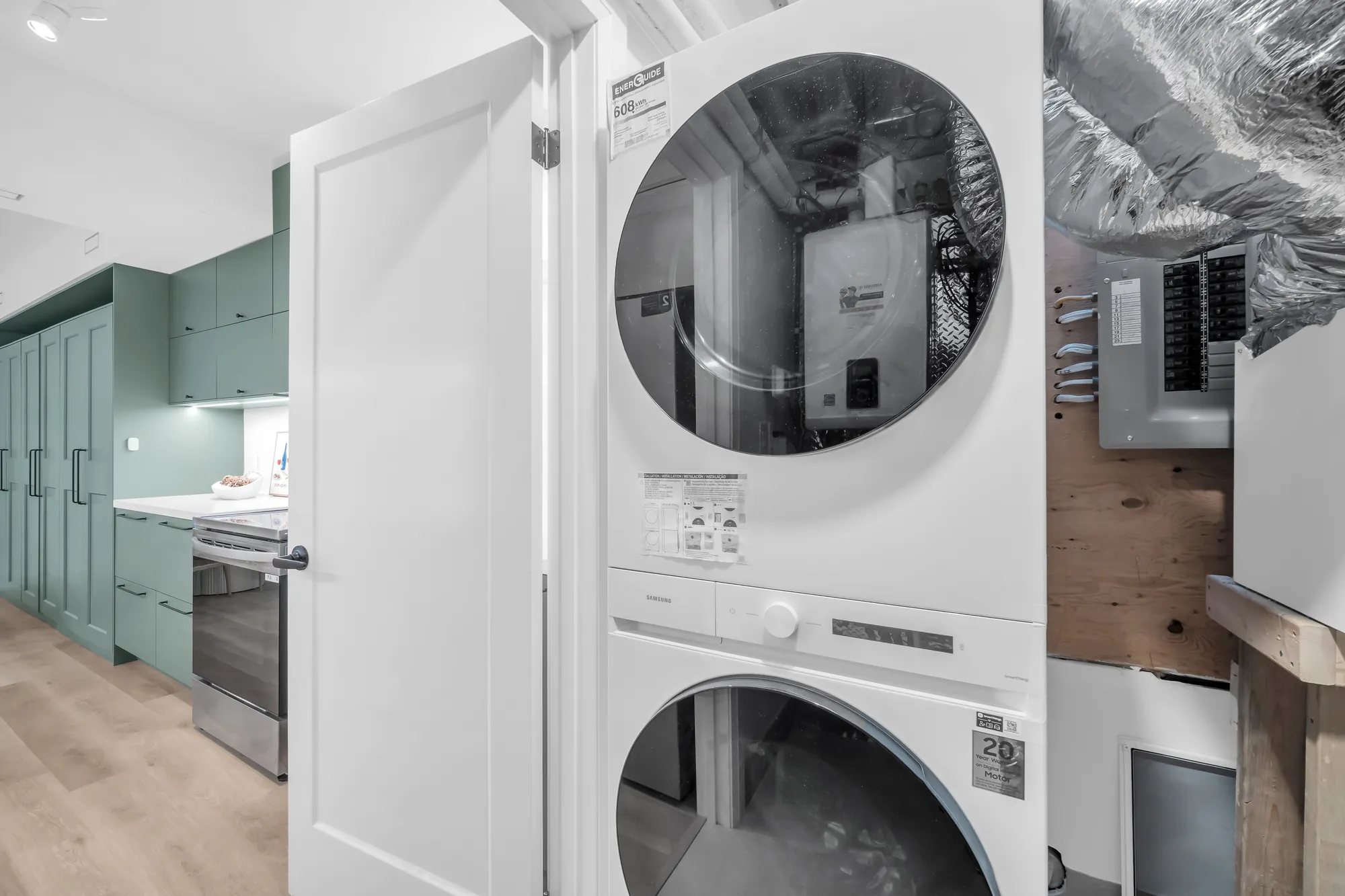 The lower level's laundry: a stacked washer and dryer standing in the mechanical room, ducting, a wall-hung heating unit and the electrical panel beside them, the green kitchen through the door